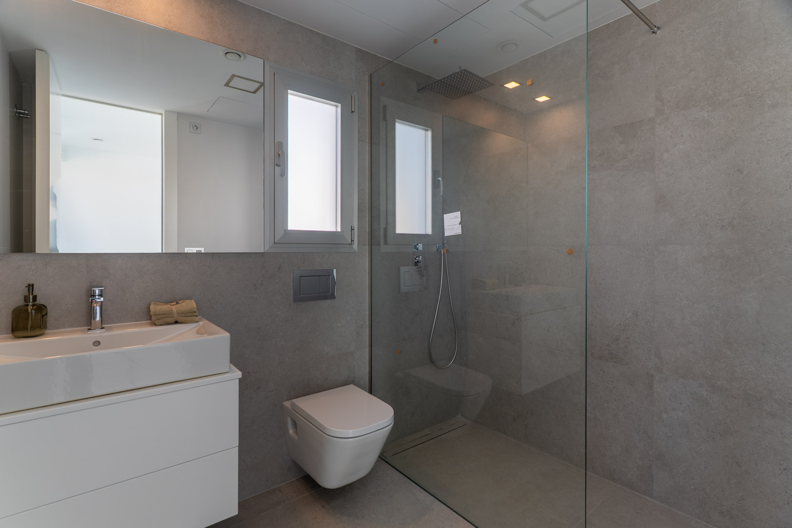 Contemporary bathroom with grey tiling, walk-in shower with glass screen, wall-mounted toilet, and integrated sink on a minimalist white vanity.