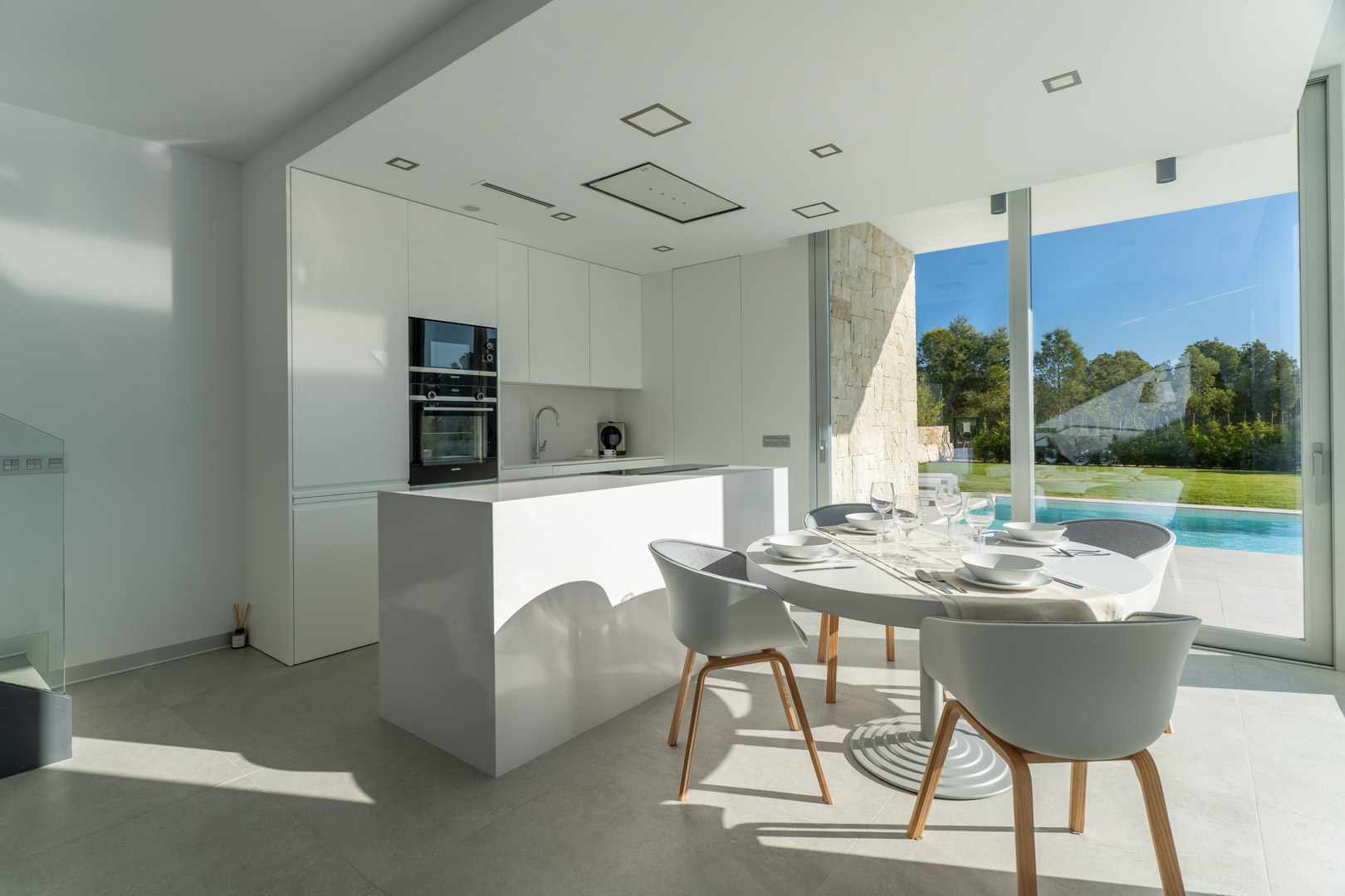 Minimalist white open-plan kitchen featuring a central island and dining area, with floor-to-ceiling windows overlooking the private pool and garden.