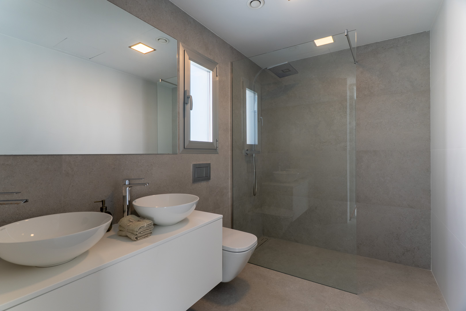 Modern bathroom featuring grey stone-effect tiling, a white floating double vanity with vessel sinks, and a walk-in shower with a frameless glass screen.