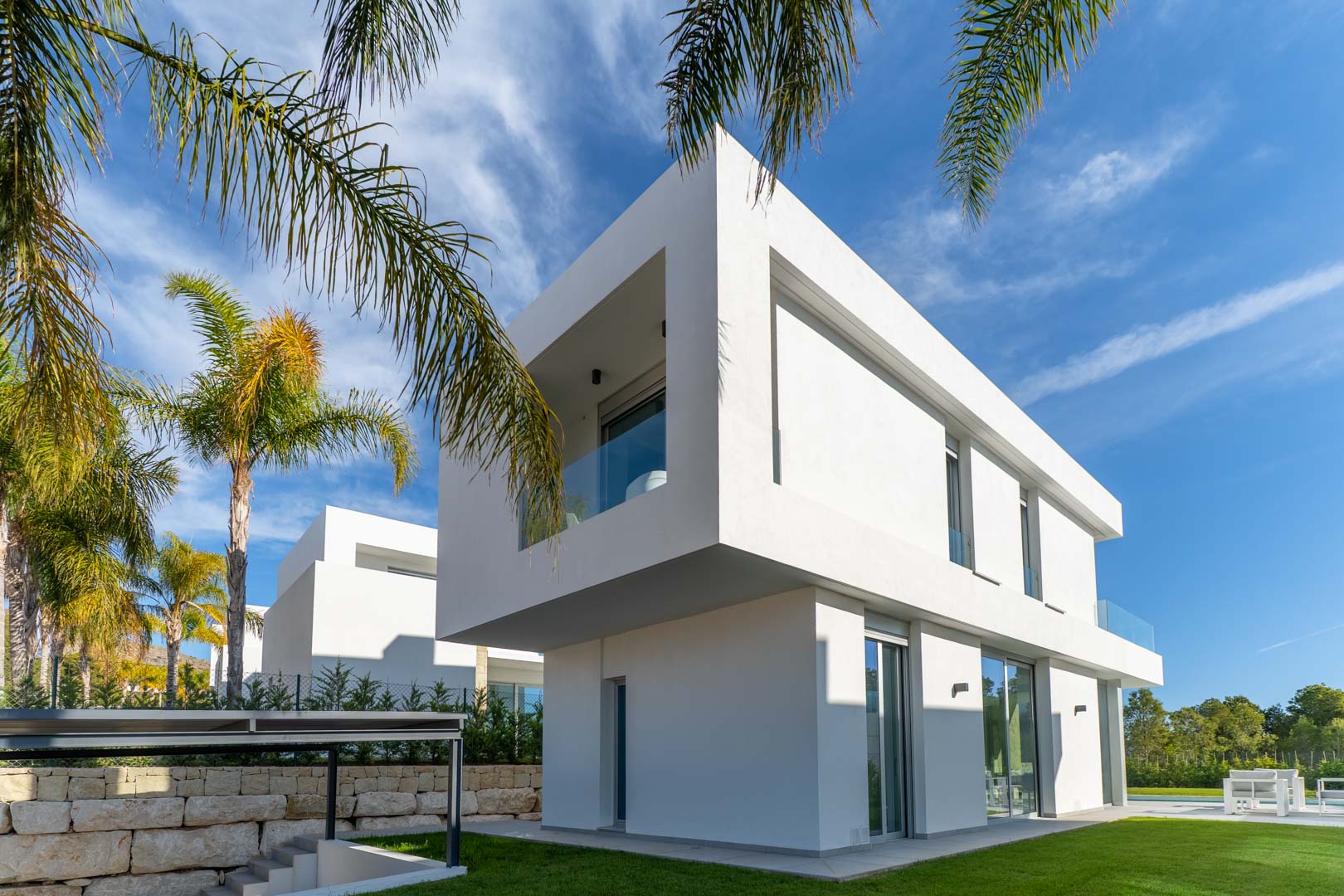 Modern two-story villa featuring a cantilevered upper floor, minimalist white facade, and large glass sliders. The exterior is framed by palm trees and a manicured lawn.