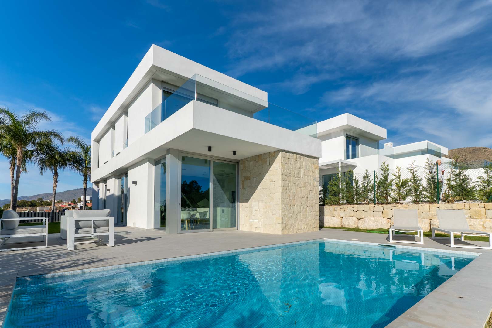 Modern villa exterior featuring a private swimming pool, minimalist white facade with natural stone accents, and glass-railed terraces.