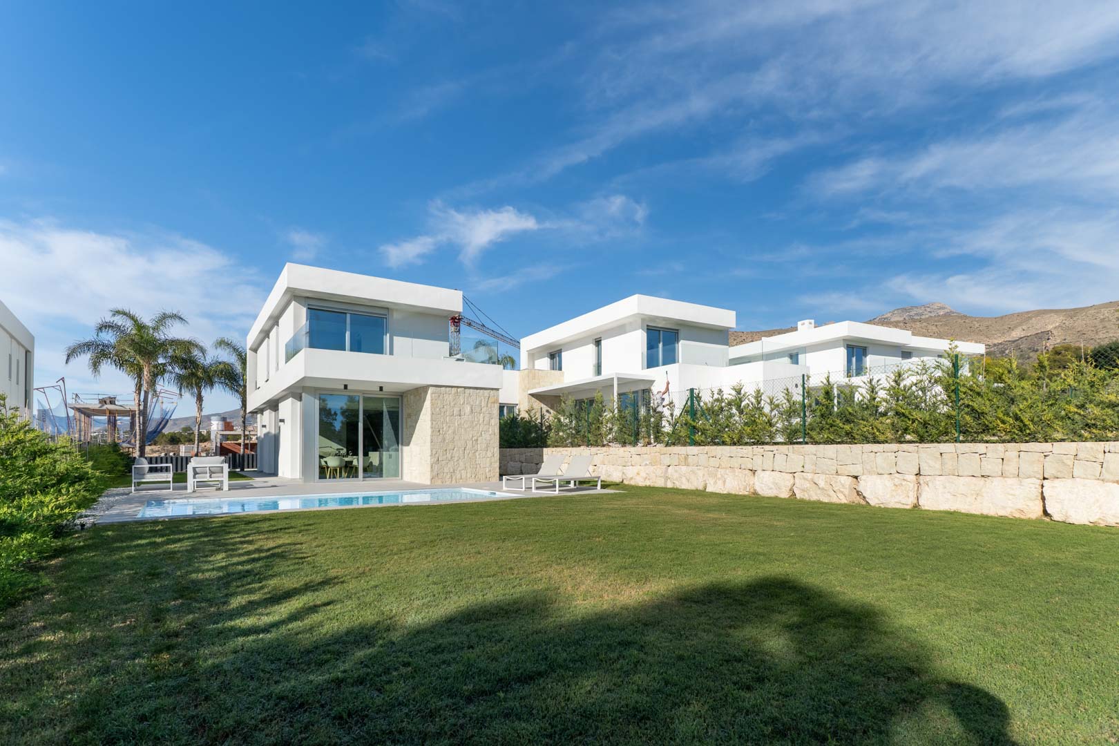 Modern villa exterior featuring a white facade with stone accents, a private swimming pool, a large lawn, and mountain views under a clear blue sky.