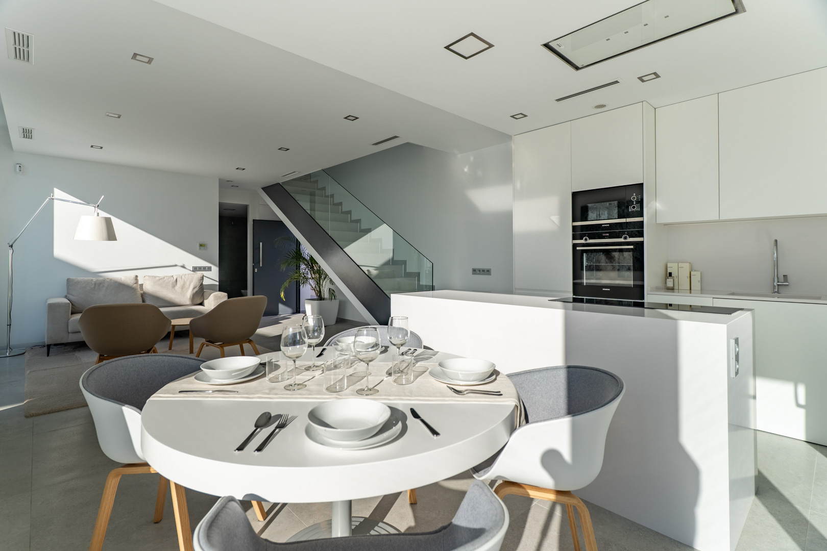 Open-plan living area featuring a minimalist white kitchen with an island, integrated appliances, and a modern staircase with a glass balustrade and metal frame.