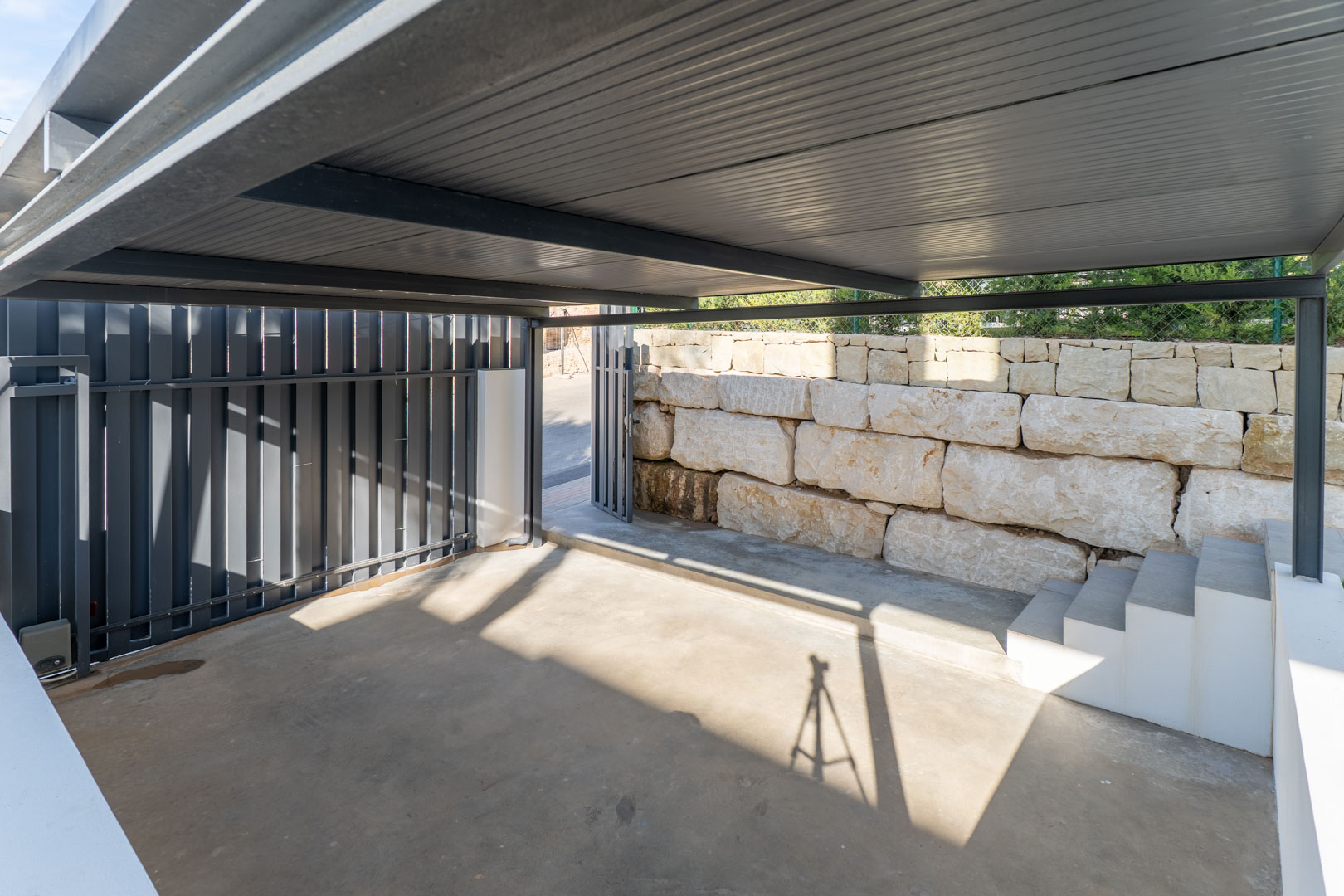 Private covered parking featuring a durable metal structure, concrete flooring, and a natural stone retaining wall with integrated access steps.