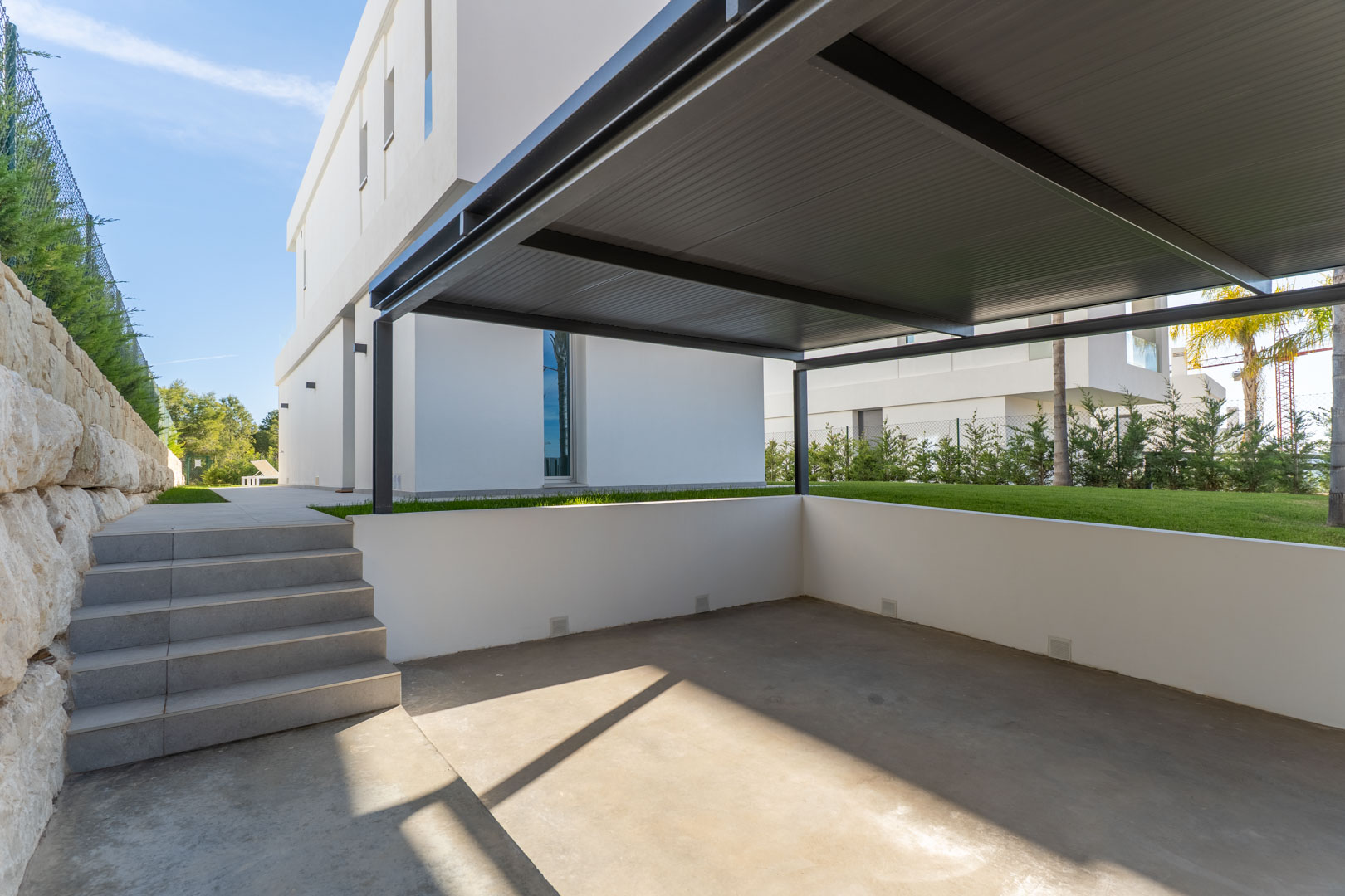 Private parking area featuring a dark metal carport, concrete flooring, and tiled steps leading to the main garden and villa entrance.