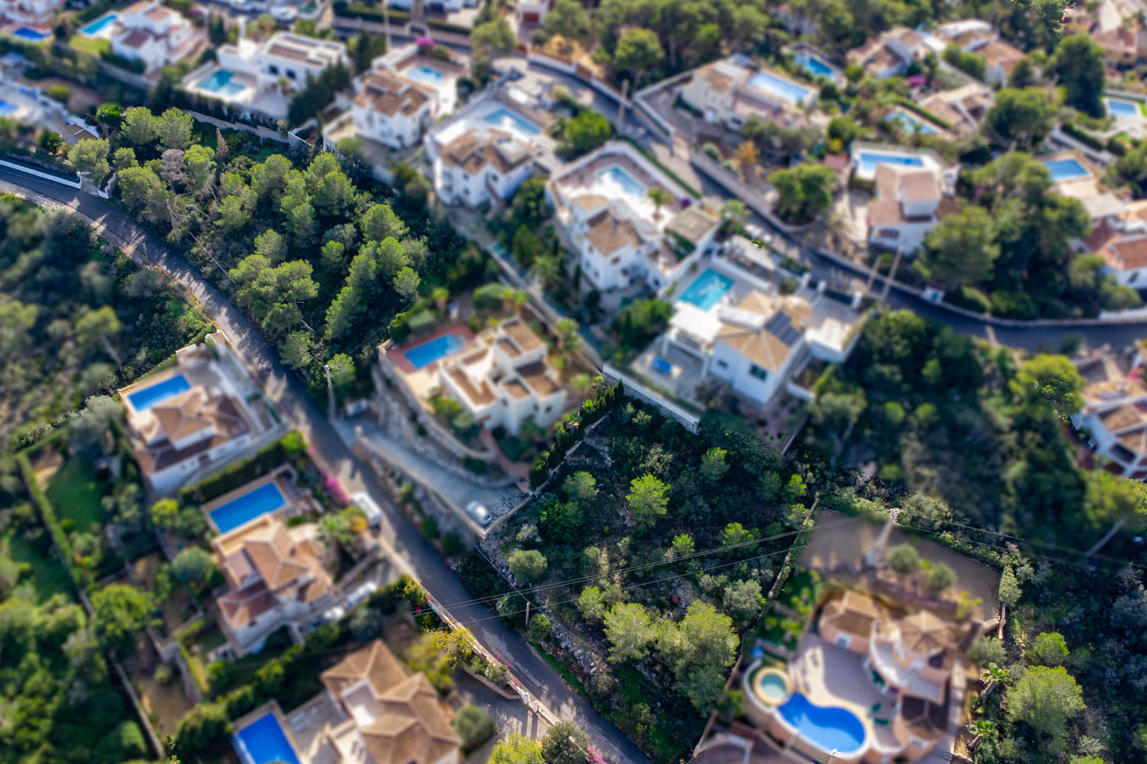 Aerial view of the sloped building plot in Granadella-Ambolo, nestled among established villas with private pools and mature pine trees.