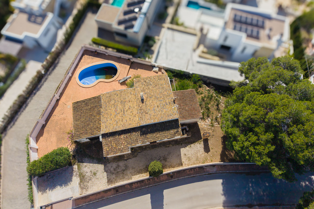Aerial view of the villa featuring its terracotta tile roof, organic-shaped swimming pool, and flat plot within a quiet residential neighborhood.