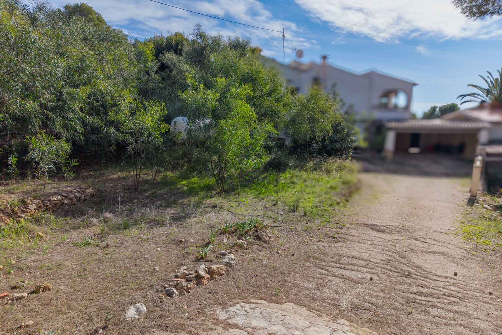 Access path to the urban plot featuring natural stone borders and Mediterranean shrubbery, with an existing villa visible in the background.