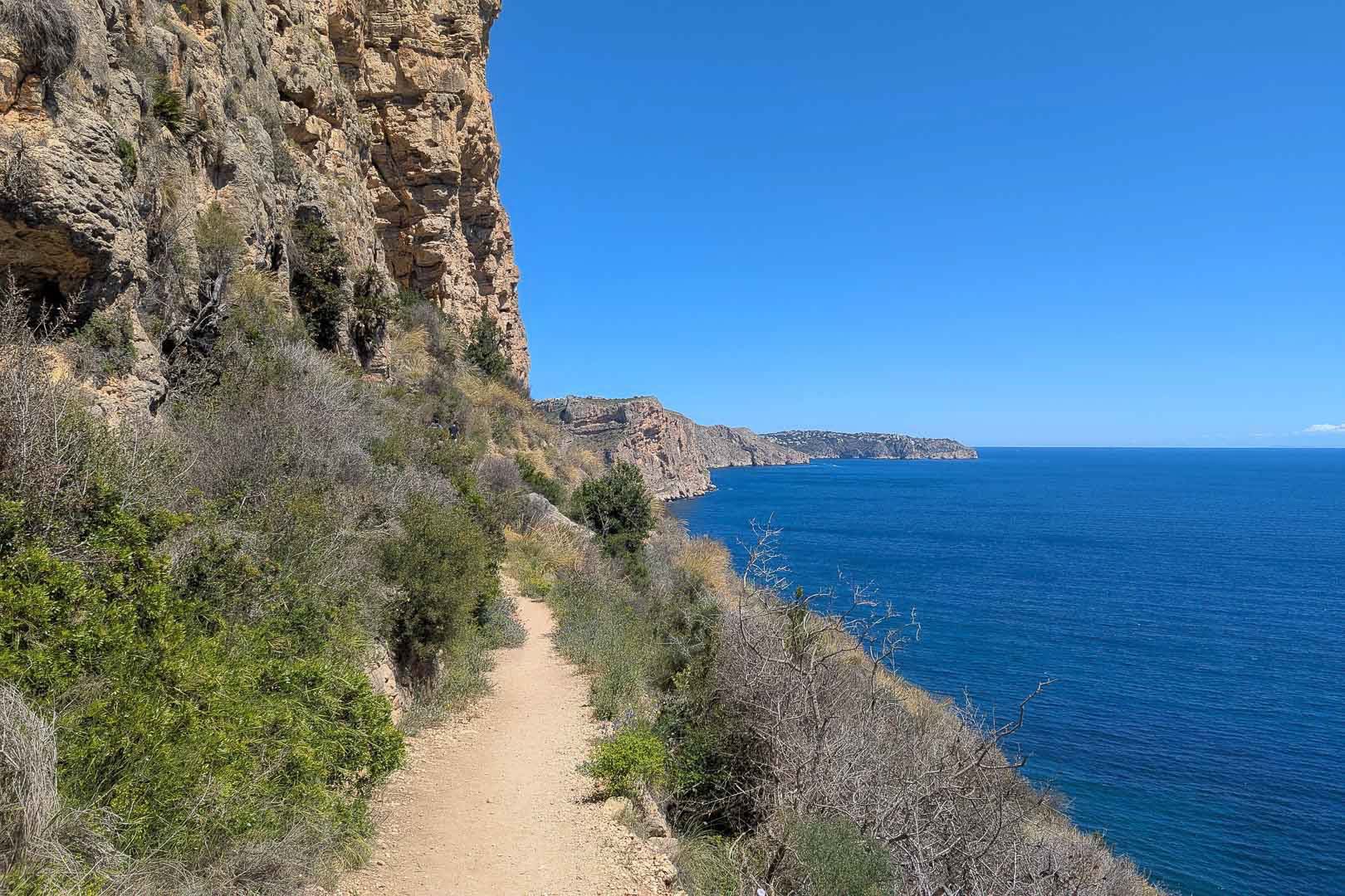 Coastal dirt path along limestone cliffs offering panoramic views of the Mediterranean Sea and native vegetation under a clear blue sky.