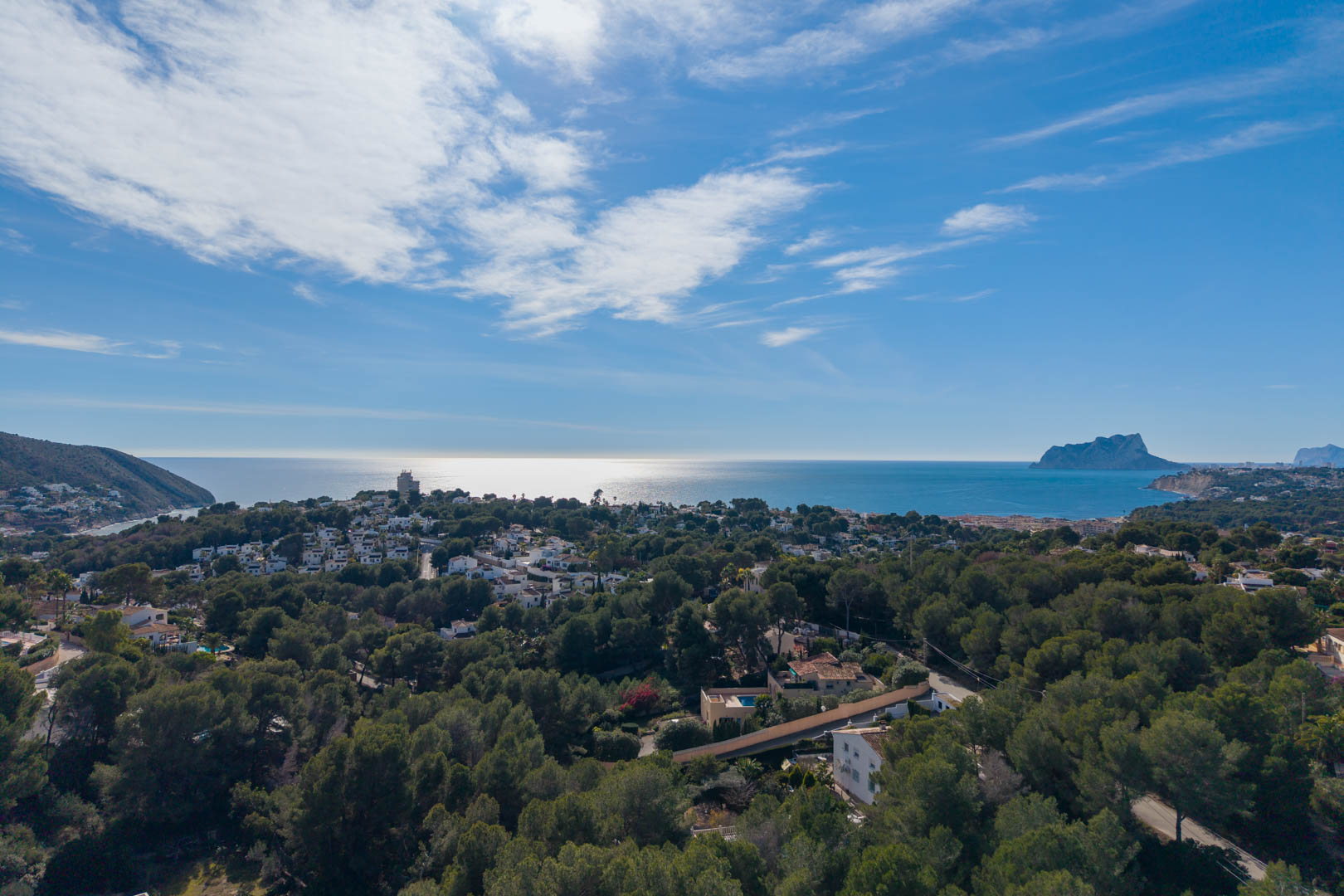 Panoramic aerial view of the Moraira coastline, featuring dense pine groves, residential villas, and the Peñón de Ifach landmark on the Mediterranean horizon.