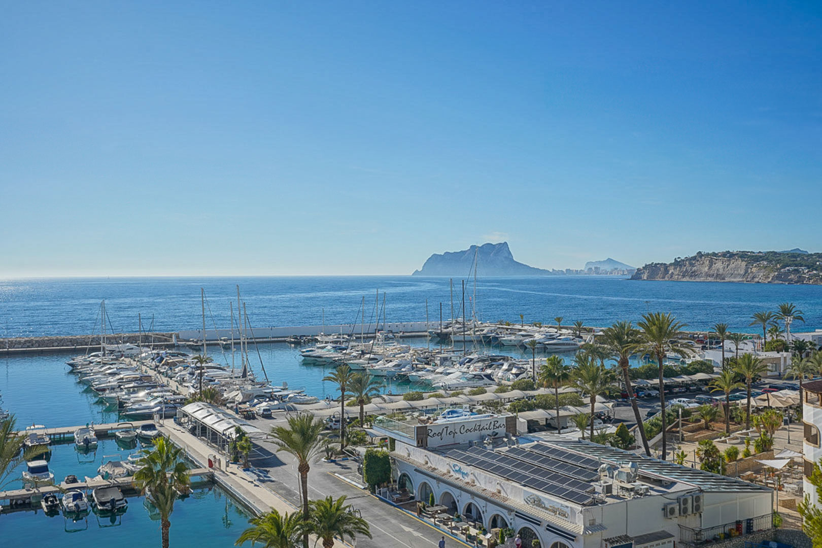 Panoramic view of Moraira's marina featuring docked yachts, palm trees, and the iconic Peñón de Ifach on the horizon under a clear blue sky.