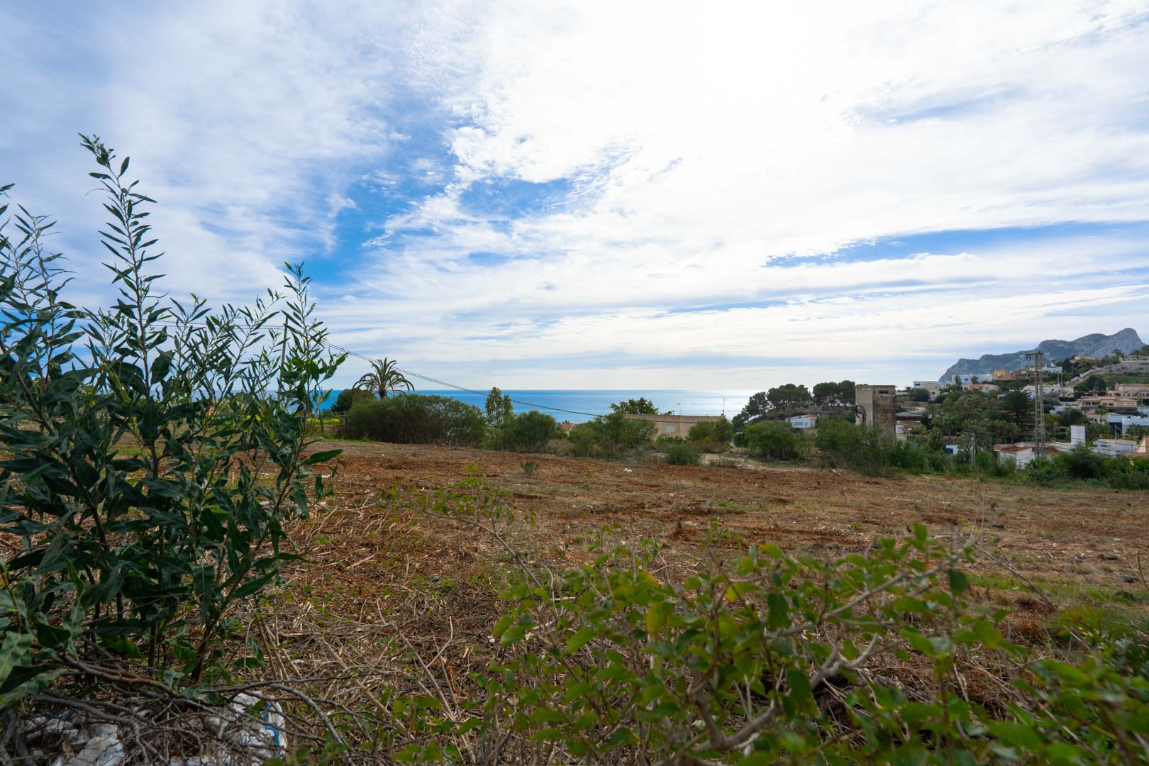 Elevated plot of land featuring panoramic views of the Mediterranean Sea and distant mountains, surrounded by coastal vegetation and residential architecture.