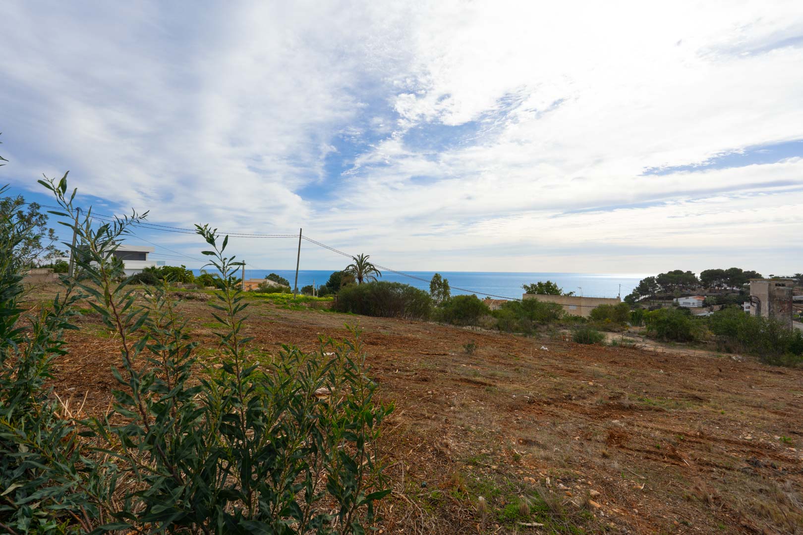 Elevated plot of land with panoramic sea views, featuring cleared earth and native Mediterranean vegetation under a bright, partly cloudy sky.