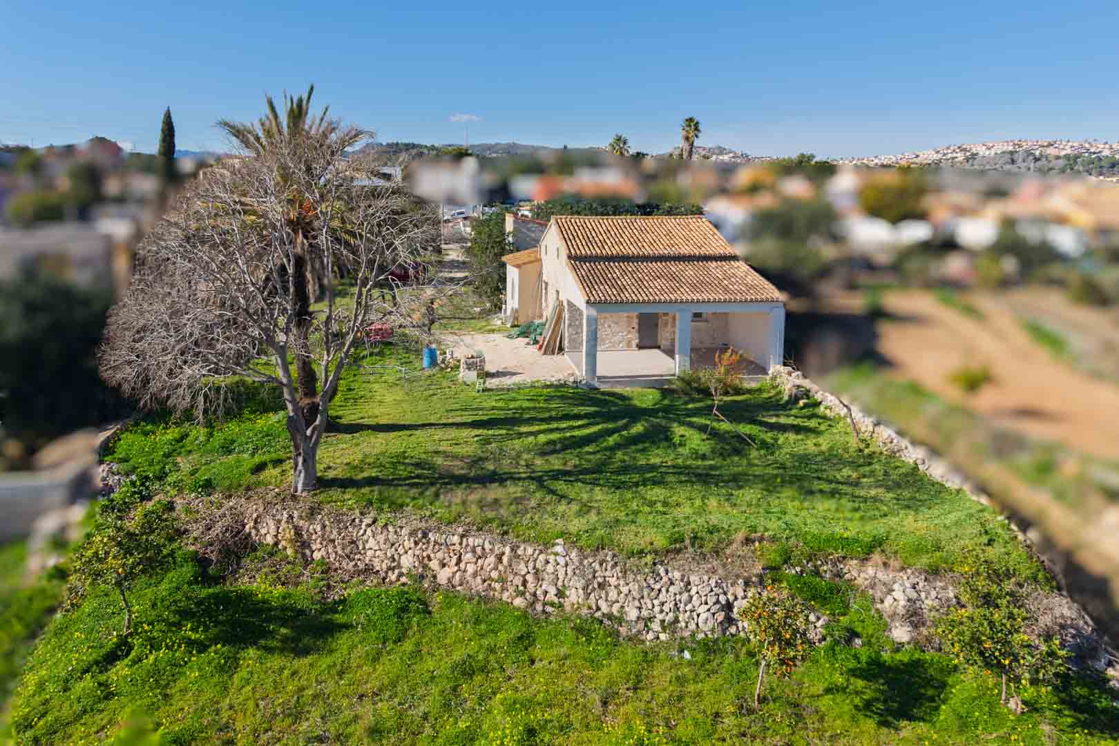 Elevated view of the traditional finca featuring a renovated terracotta roof, dry stone walls, and a spacious naya set within a green plot with fruit trees.