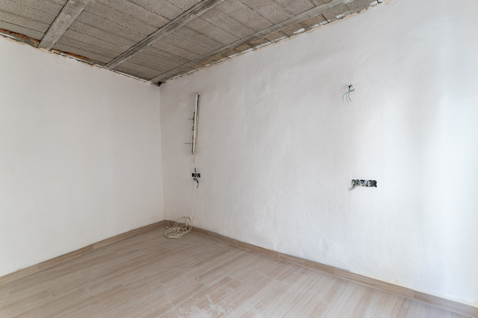 Interior room under renovation featuring wood-effect ceramic flooring, white plastered walls, and an exposed concrete joist ceiling with electrical wiring.