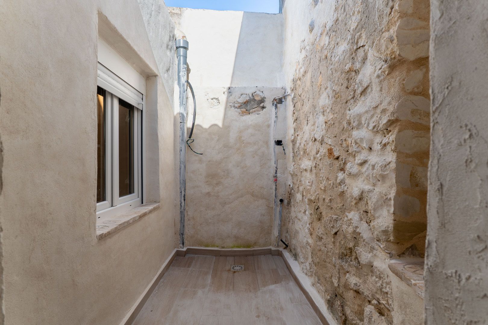 Narrow interior patio featuring a rustic dry stone wall, ceramic tiled flooring with drainage, and a renovated facade with a white PVC window.