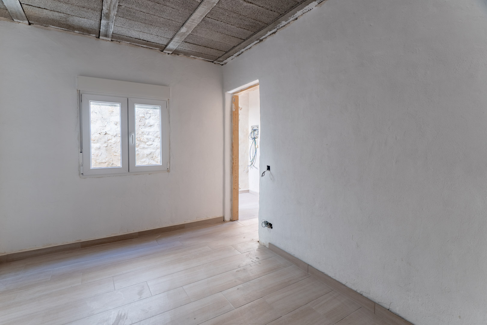 Renovated interior space featuring wood-effect ceramic flooring, white walls, and a new PVC window. The ceiling displays the structural concrete joists.