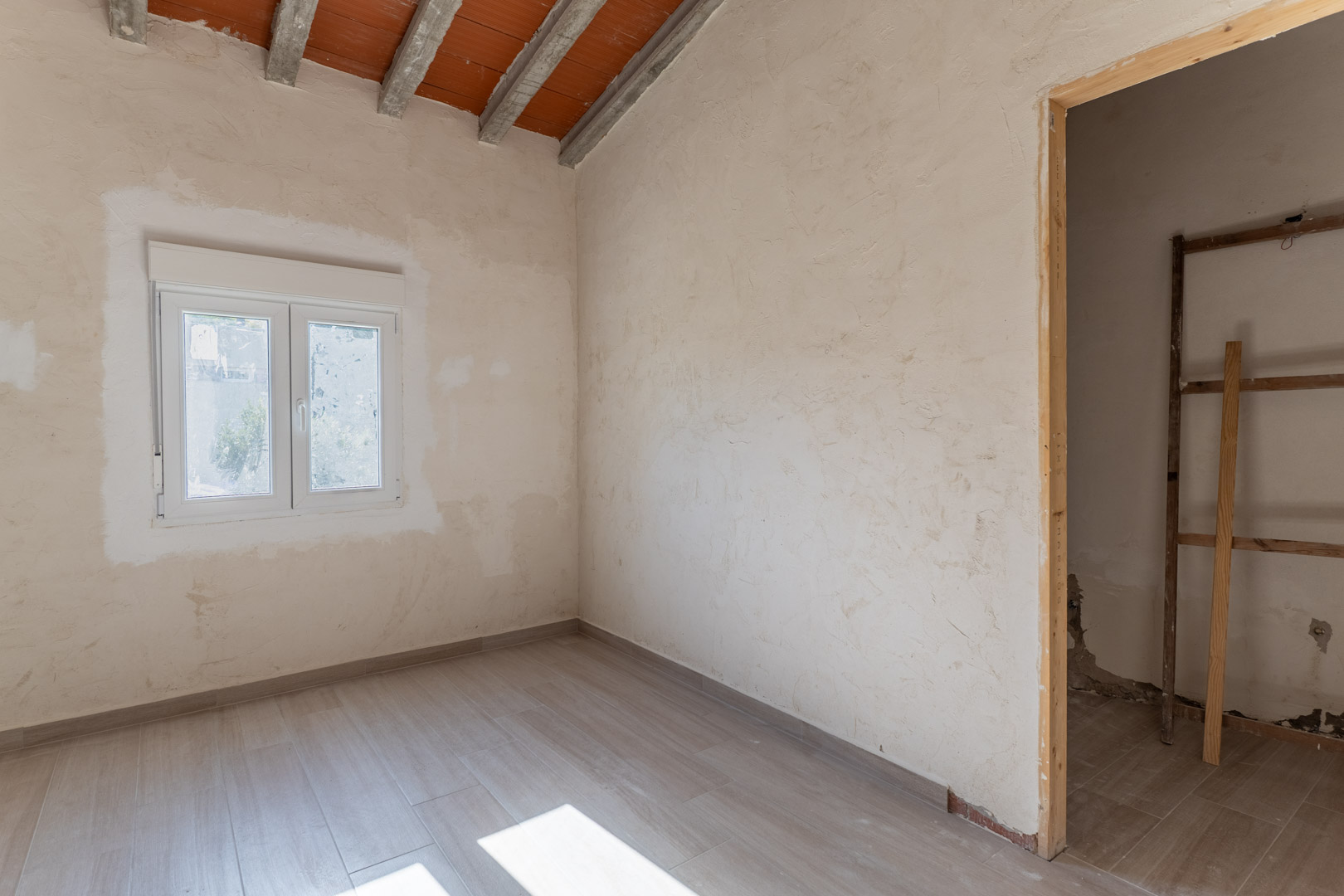 Renovated room featuring wood-effect ceramic flooring, a white PVC window, and a sloped ceiling with exposed concrete beams and terracotta tiles.