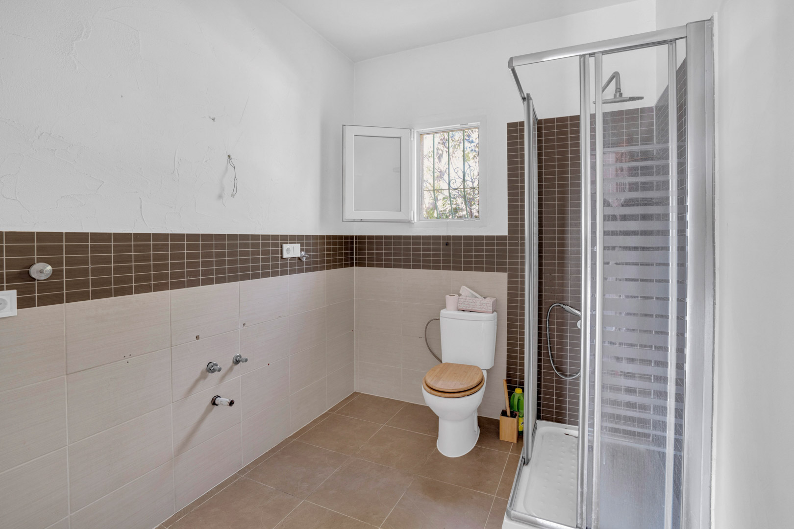 Bathroom featuring a walk-in shower with glass enclosure, brown mosaic tiling, and a toilet with a wooden lid. The walls are finished with neutral ceramic tiles and a small window.