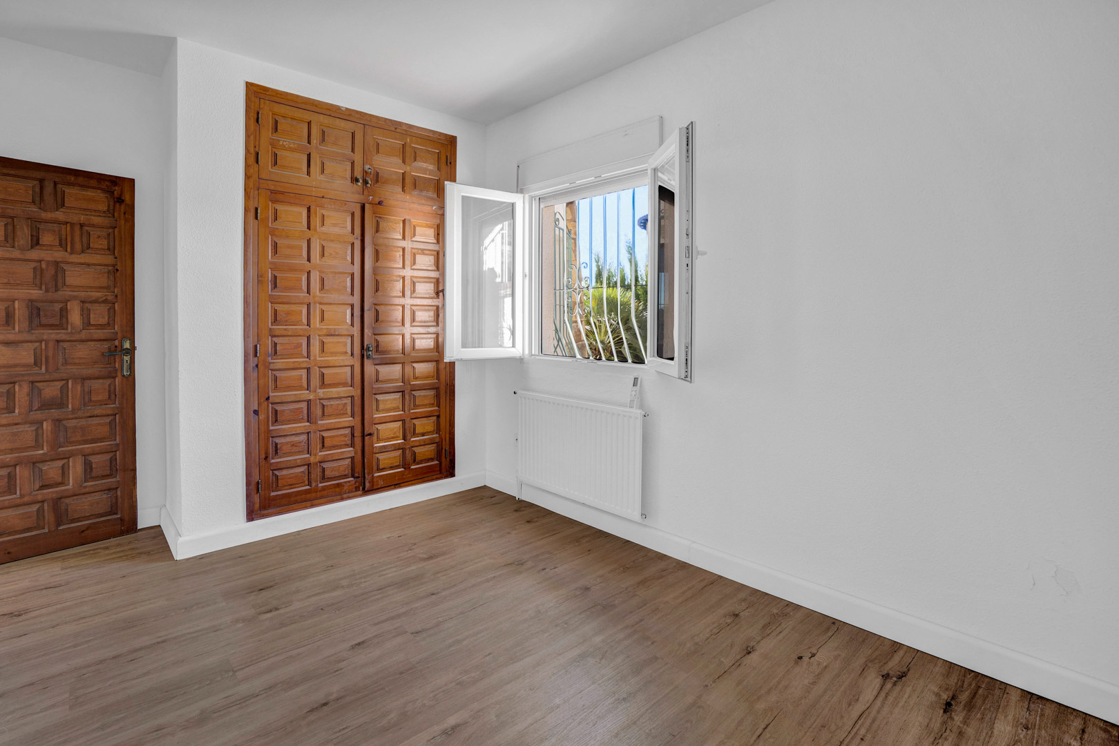 Bright bedroom featuring a built-in carved wood wardrobe, laminate flooring, gas radiator, and a white PVC window overlooking the garden.