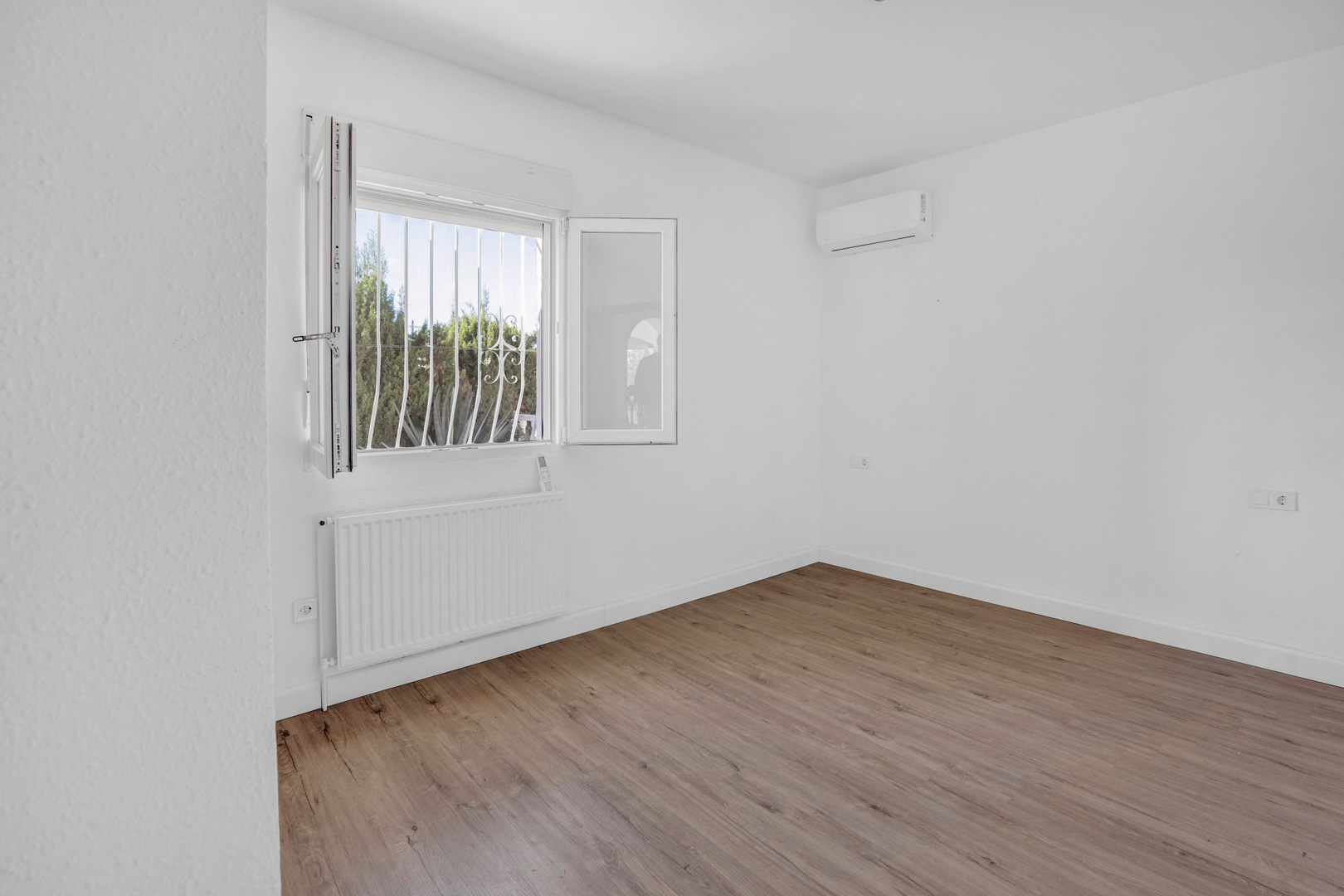 Bright bedroom featuring wood-effect laminate flooring, equipped with a central heating radiator and a wall-mounted split air conditioning unit.