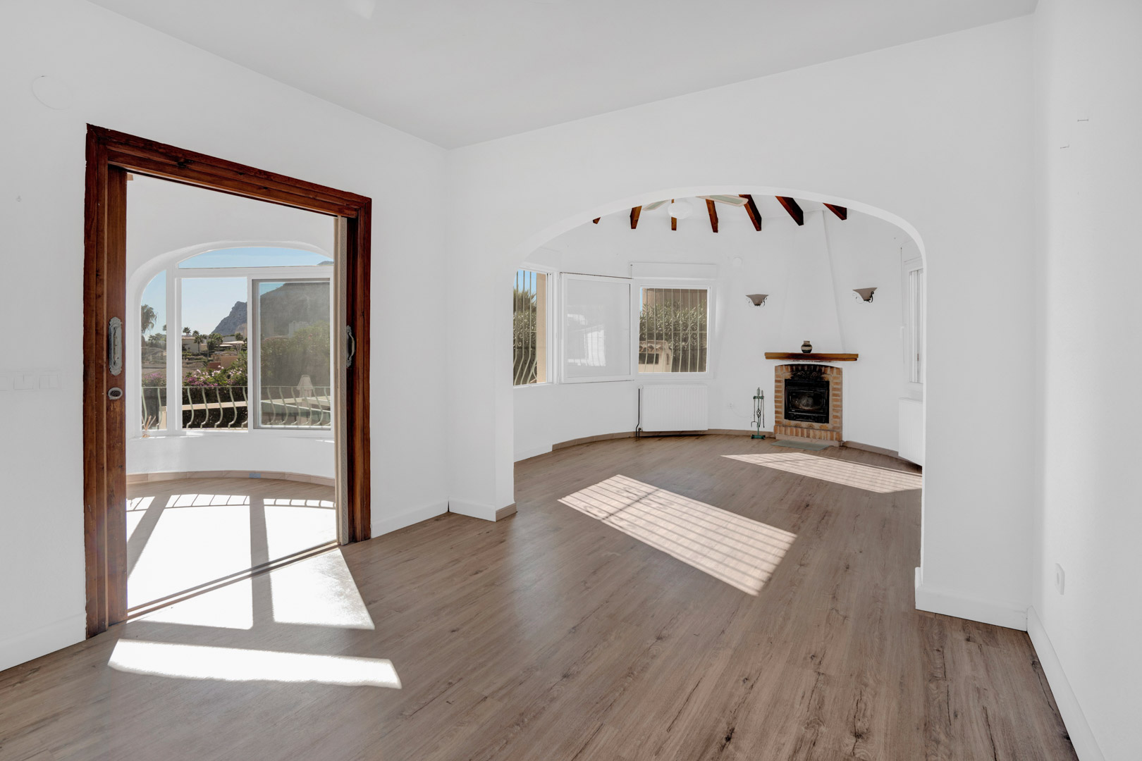 Bright living area featuring a brick fireplace, exposed wooden beams, and a wooden door frame leading to a sunroom with views of the Peñón de Ifach.