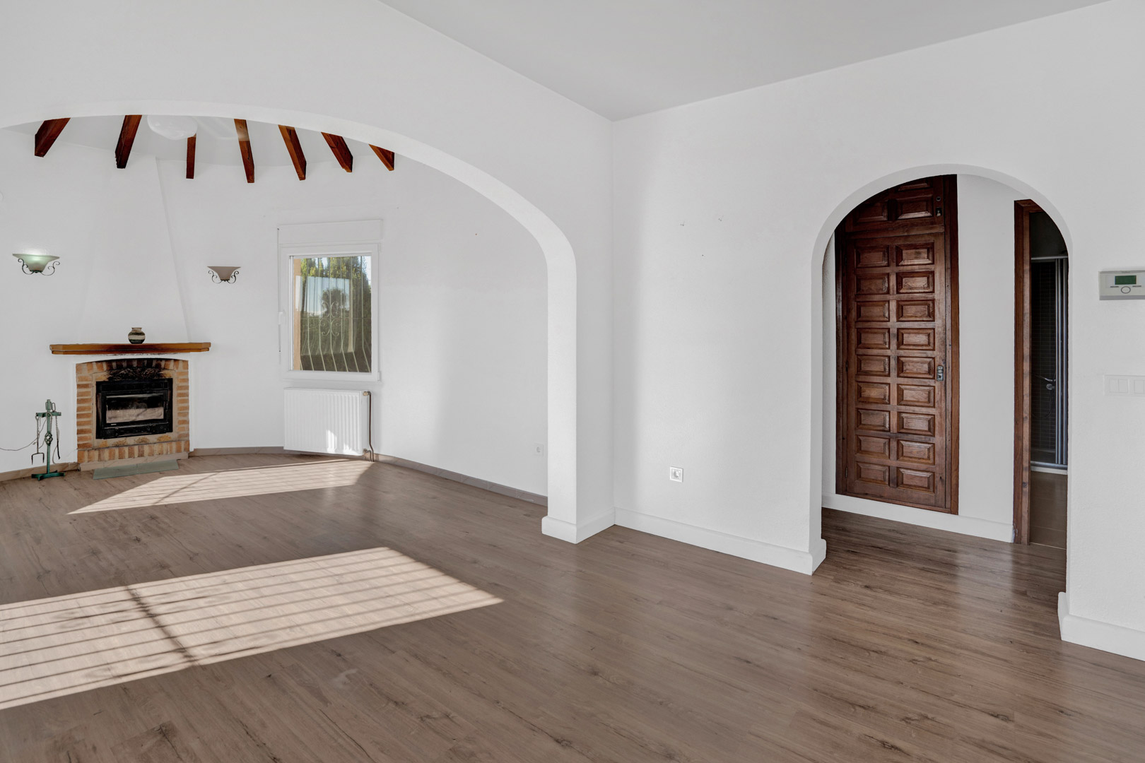 Bright living area featuring a brick fireplace, exposed wooden ceiling beams, and laminate flooring. Includes architectural archways, radiator heating, and a solid wood paneled door.