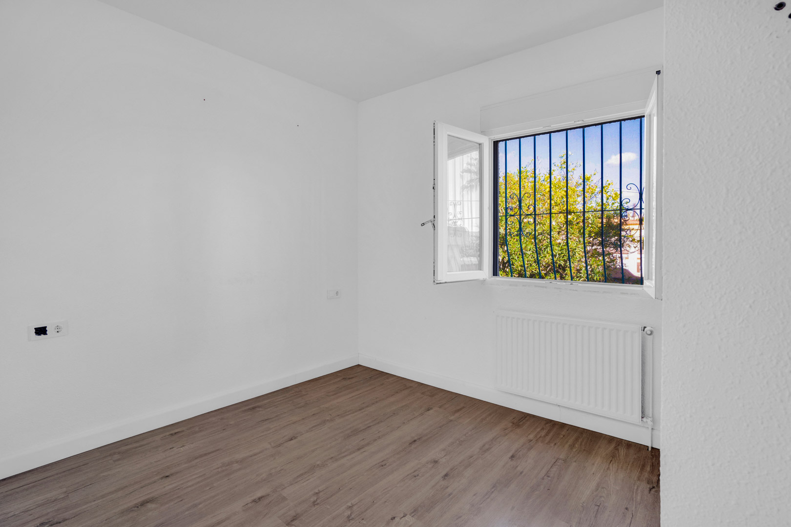 Bright room featuring wood-effect flooring, white walls, and a window with decorative blue security bars overlooking the garden, equipped with a radiator.