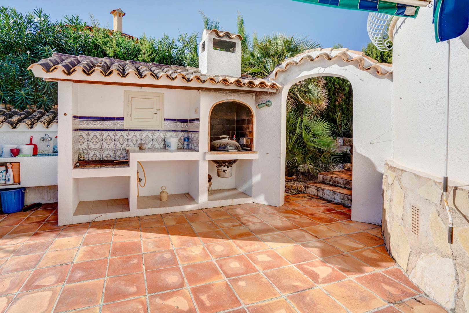 Built-in outdoor barbecue area featuring traditional tile work, a prep counter, and terracotta flooring, adjacent to an arched Mediterranean garden entrance.
