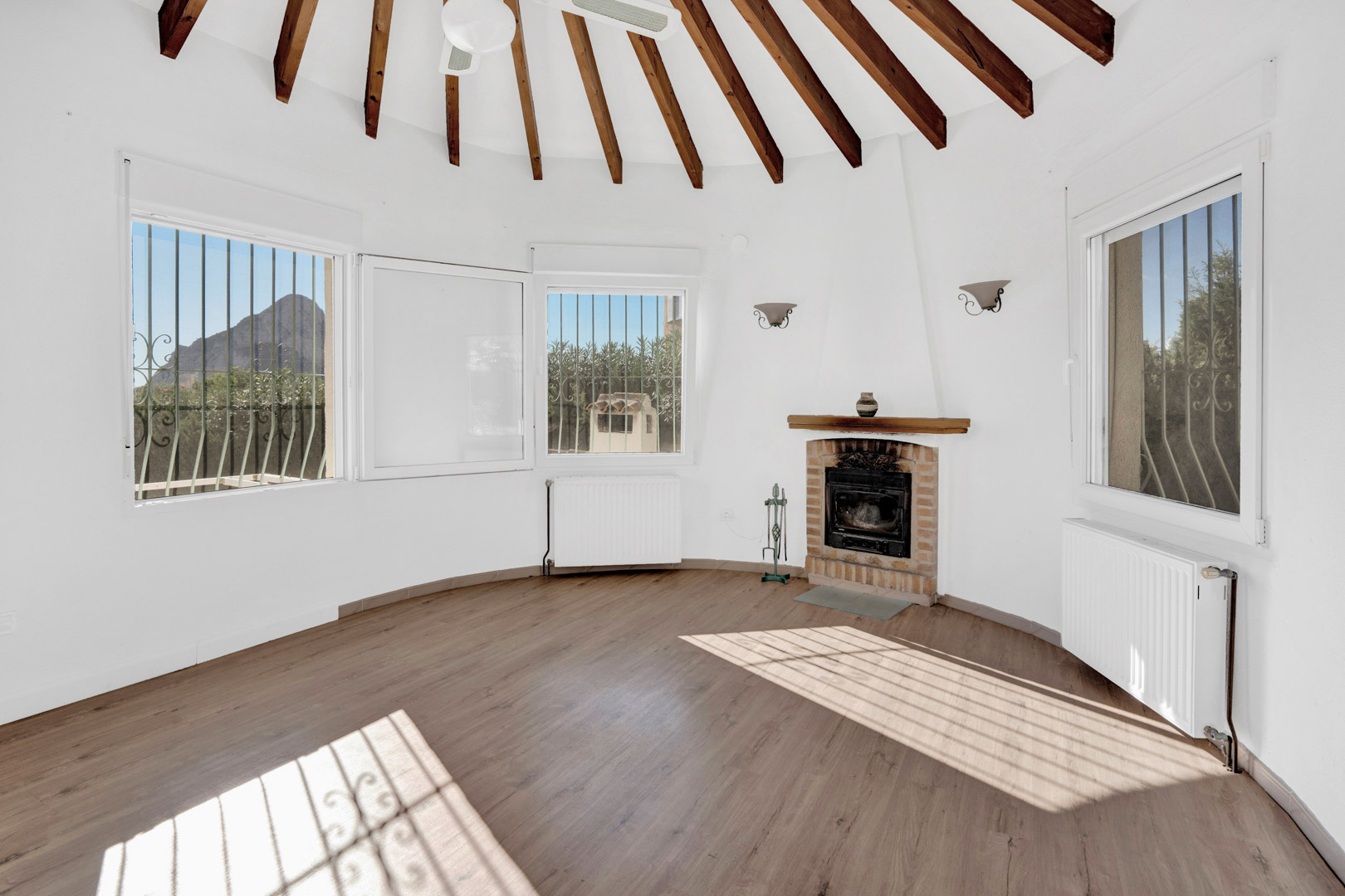 Circular living area featuring a radial beamed ceiling, brick fireplace, and large windows offering views of the Peñón de Ifach.