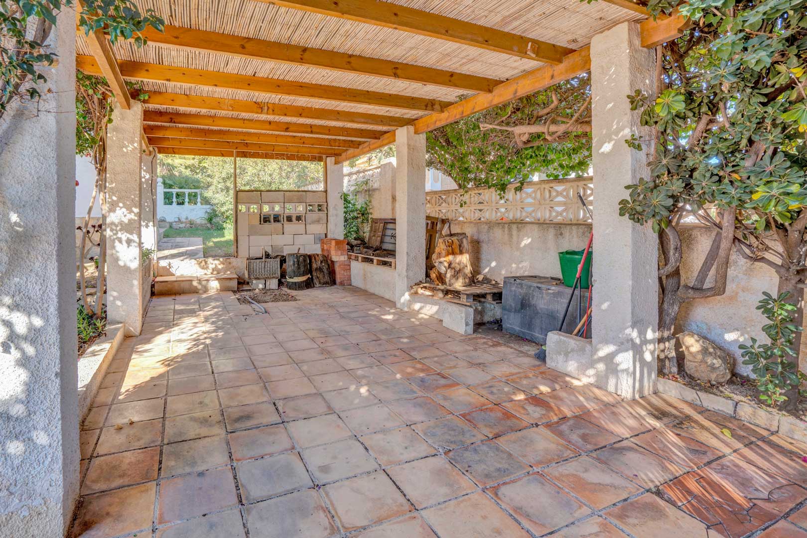 Covered carport featuring wooden beams and a reed ceiling, with terracotta-tiled flooring and a practical side area for firewood and equipment storage.