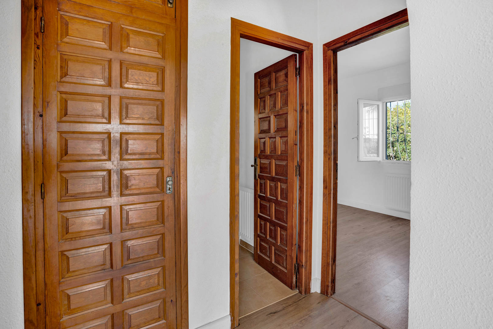 Distribution hallway featuring traditional paneled wood doors, white textured walls, and laminate flooring leading to bright bedrooms with exterior views.