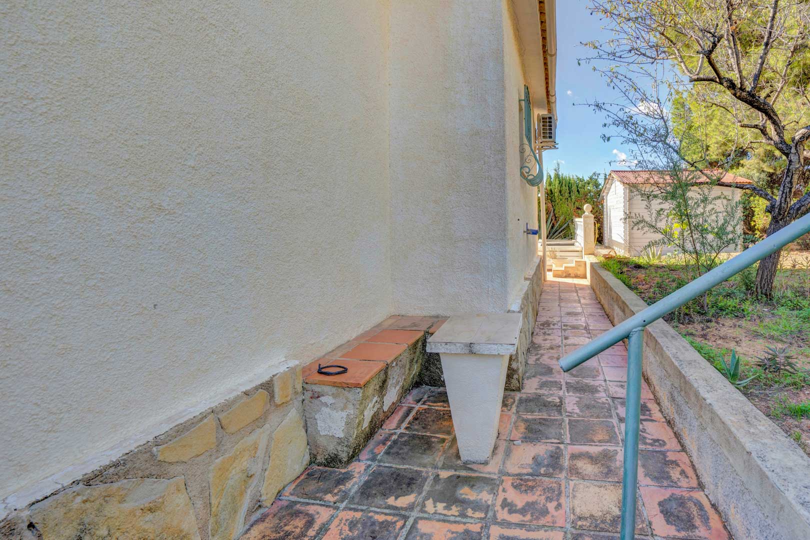 Exterior side walkway featuring terracotta tile flooring, a natural stone base, and access to the garden area with a storage shed.