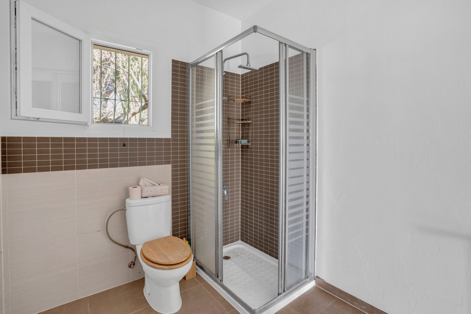 Functional bathroom featuring a corner shower with brown mosaic tiles, glass enclosure, toilet with wooden lid, and a window providing natural light.