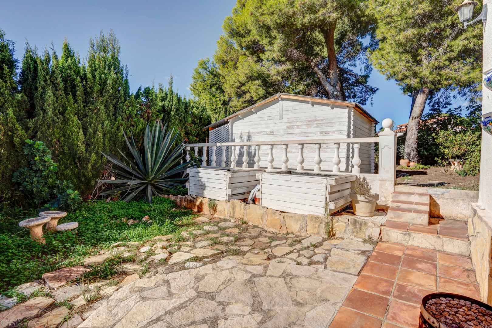 Garden area featuring irregular stone paving, a white wooden storage shed, and mature Mediterranean vegetation including pines and a large agave.