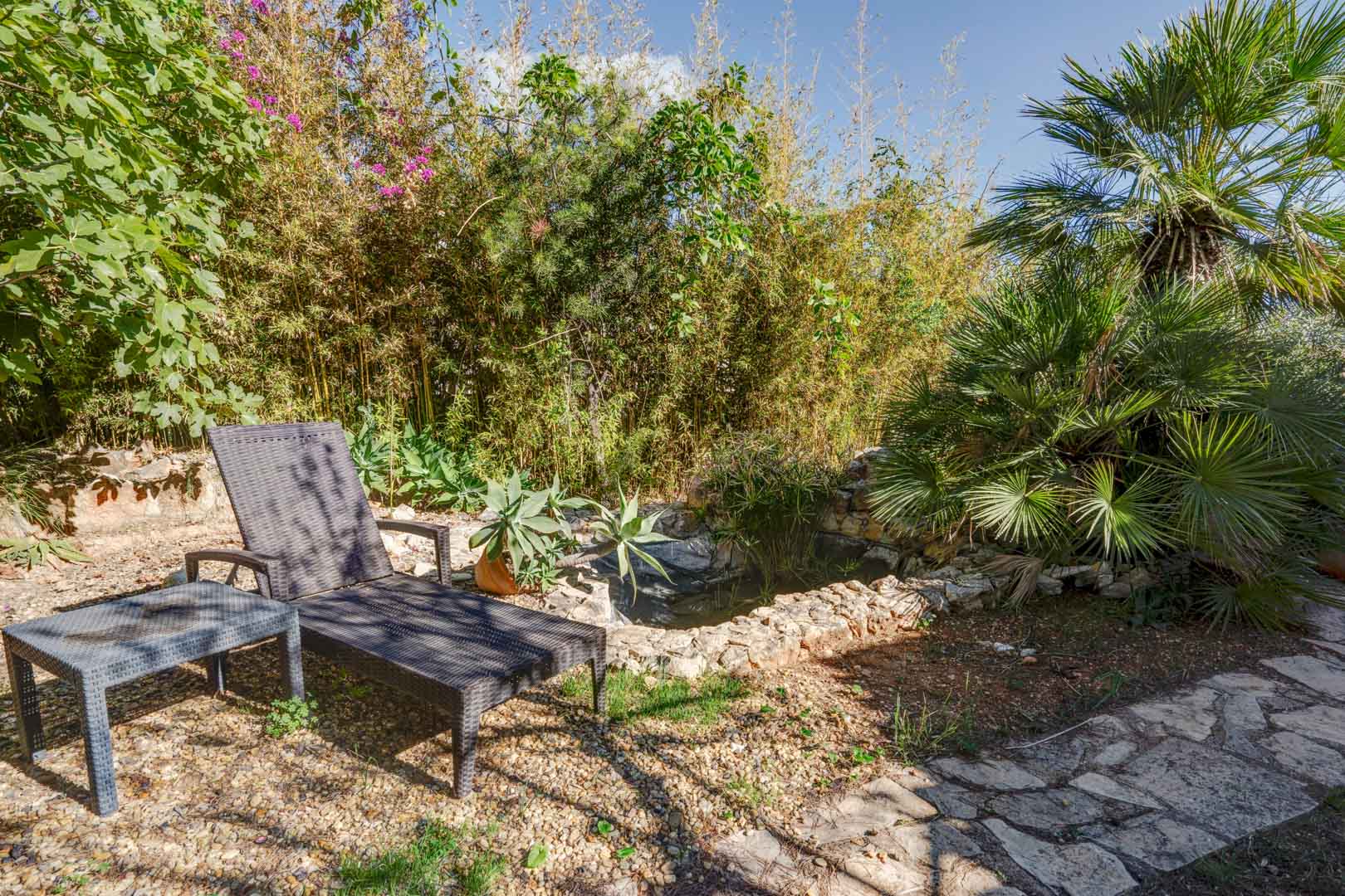Garden relaxation area featuring a synthetic rattan sun lounger and side table next to a stone-lined pond, surrounded by mature bamboo and palm trees.