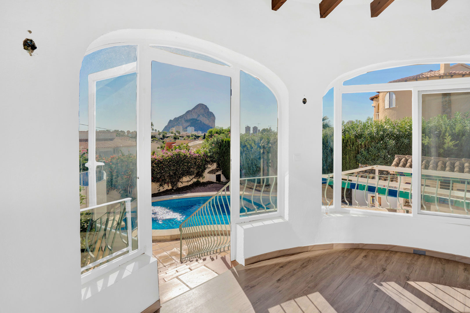 Glazed sunroom featuring wood-effect flooring, exposed ceiling beams, and arched windows with panoramic views of the Peñón de Ifach and the private pool.