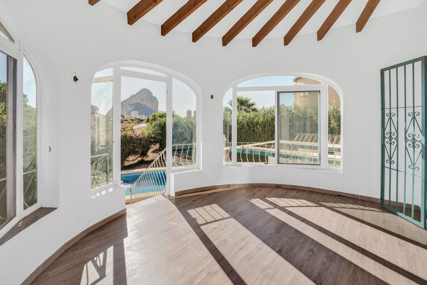 Glazed terrace featuring exposed wooden beams, laminate flooring, and arched windows with direct views of the Peñón de Ifach and the private pool.
