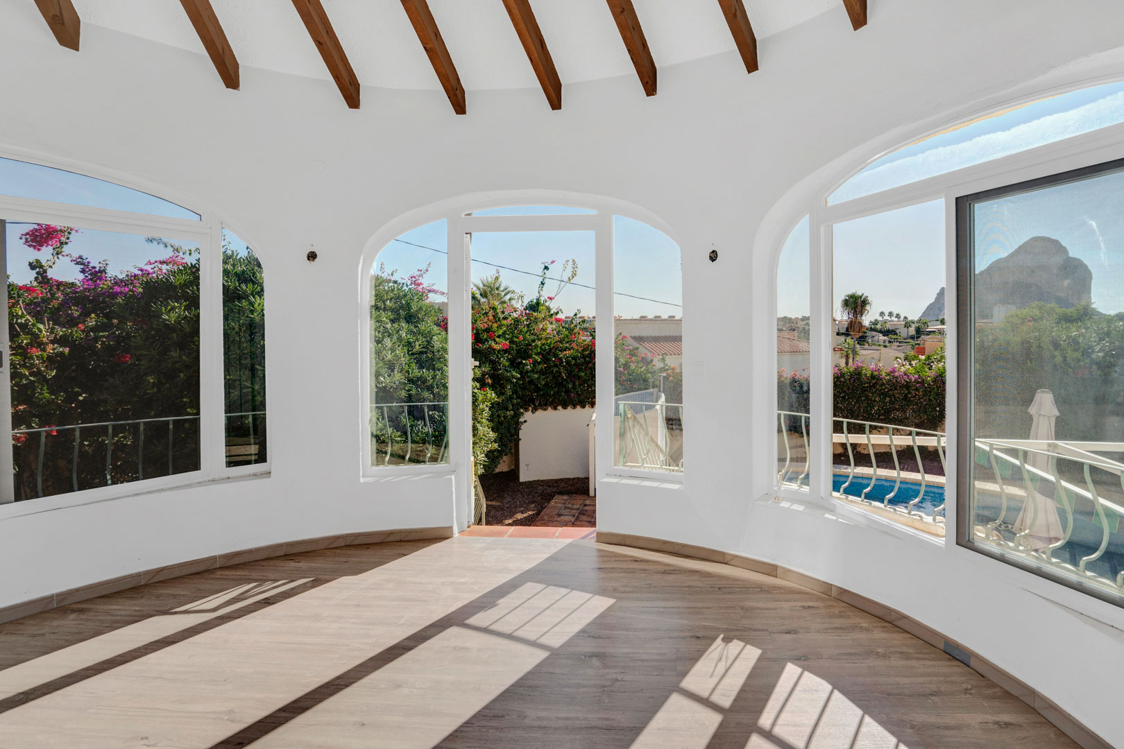 Glazed terrace featuring exposed wooden ceiling beams, laminate flooring, and arched windows with views of the Peñón de Ifach and the private pool area.