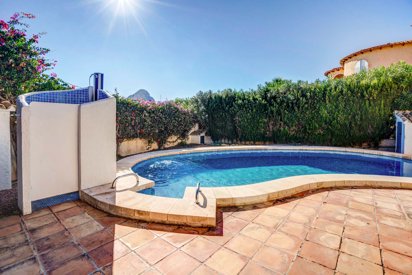 Kidney-shaped private pool featuring a terracotta terrace, outdoor shower with blue mosaic tiles, and views of the Peñón de Ifach over mature privacy hedges.