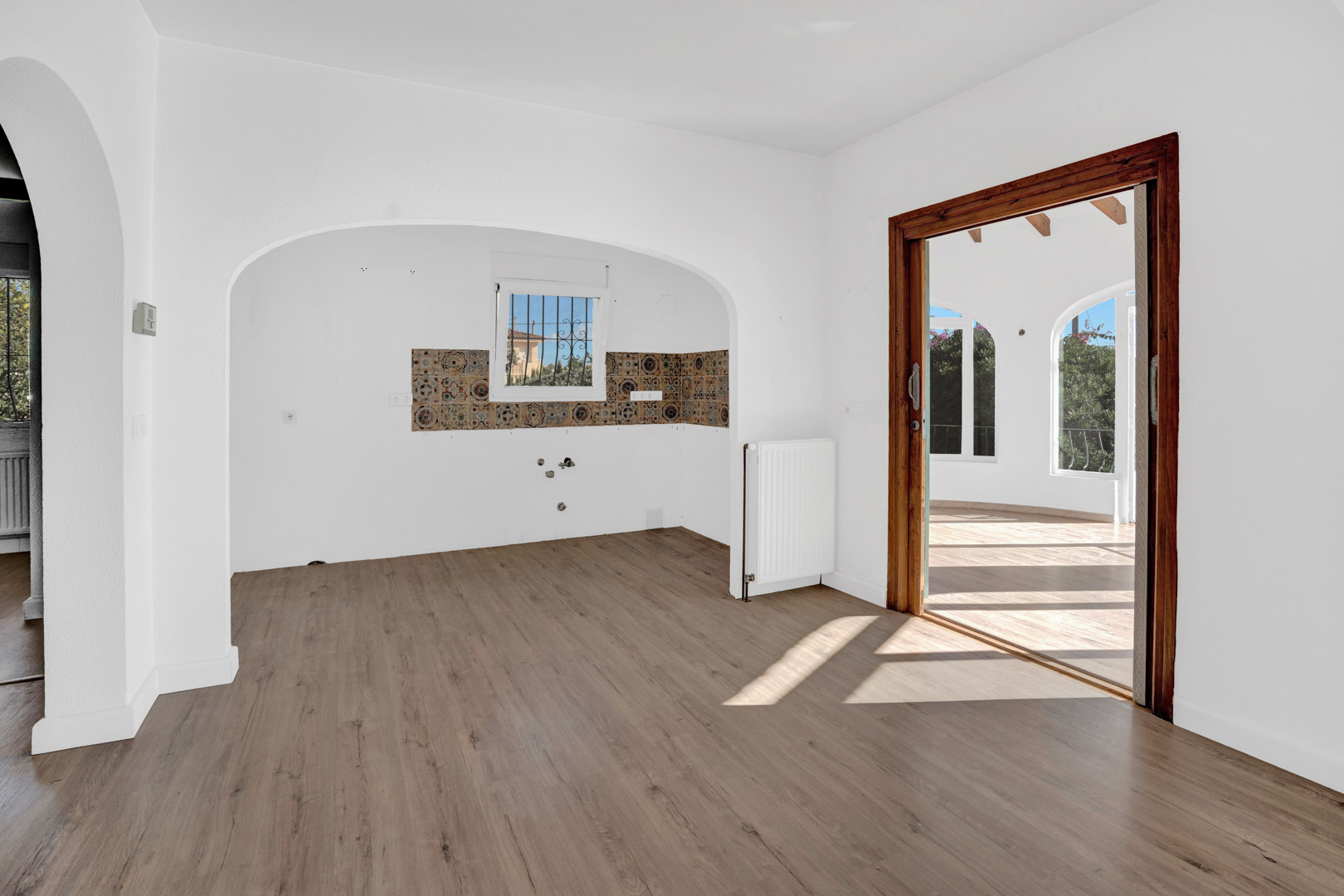 Living area featuring laminate flooring, a radiator, and an arched kitchen alcove with patterned tiles. A wooden sliding door leads to the bright glazed terrace.