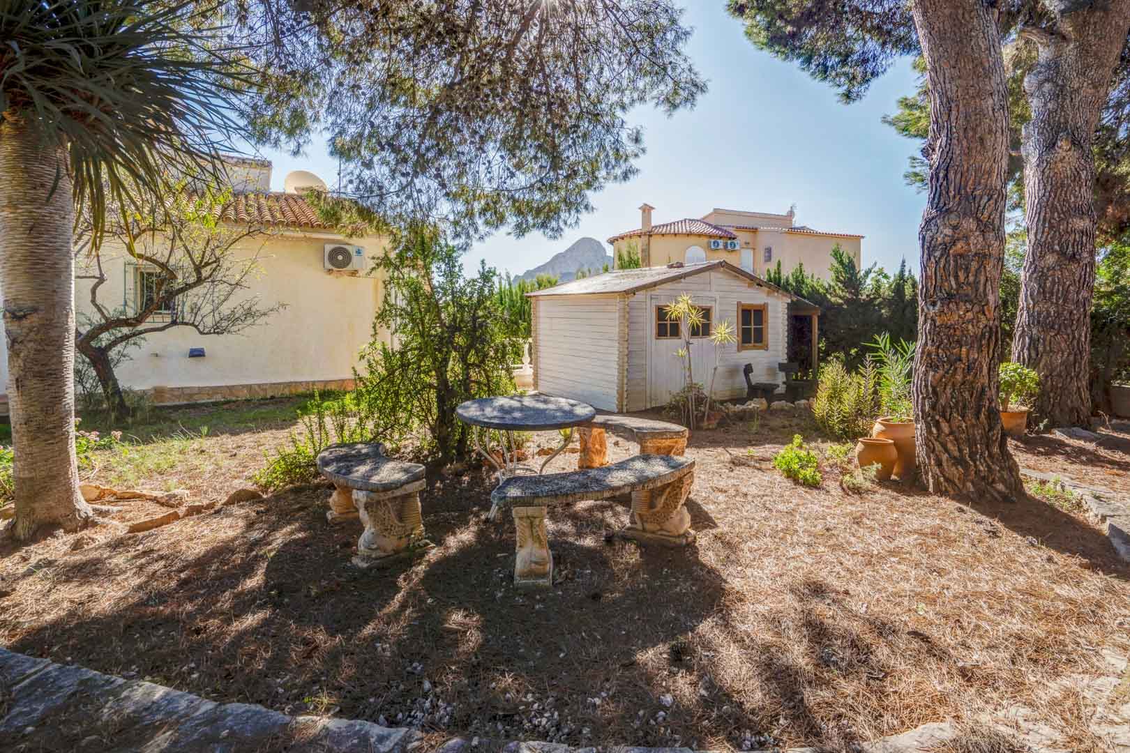 Mediterranean garden featuring a stone seating area under mature trees, a white wooden storage shed, and distant views of the Peñón de Ifach.