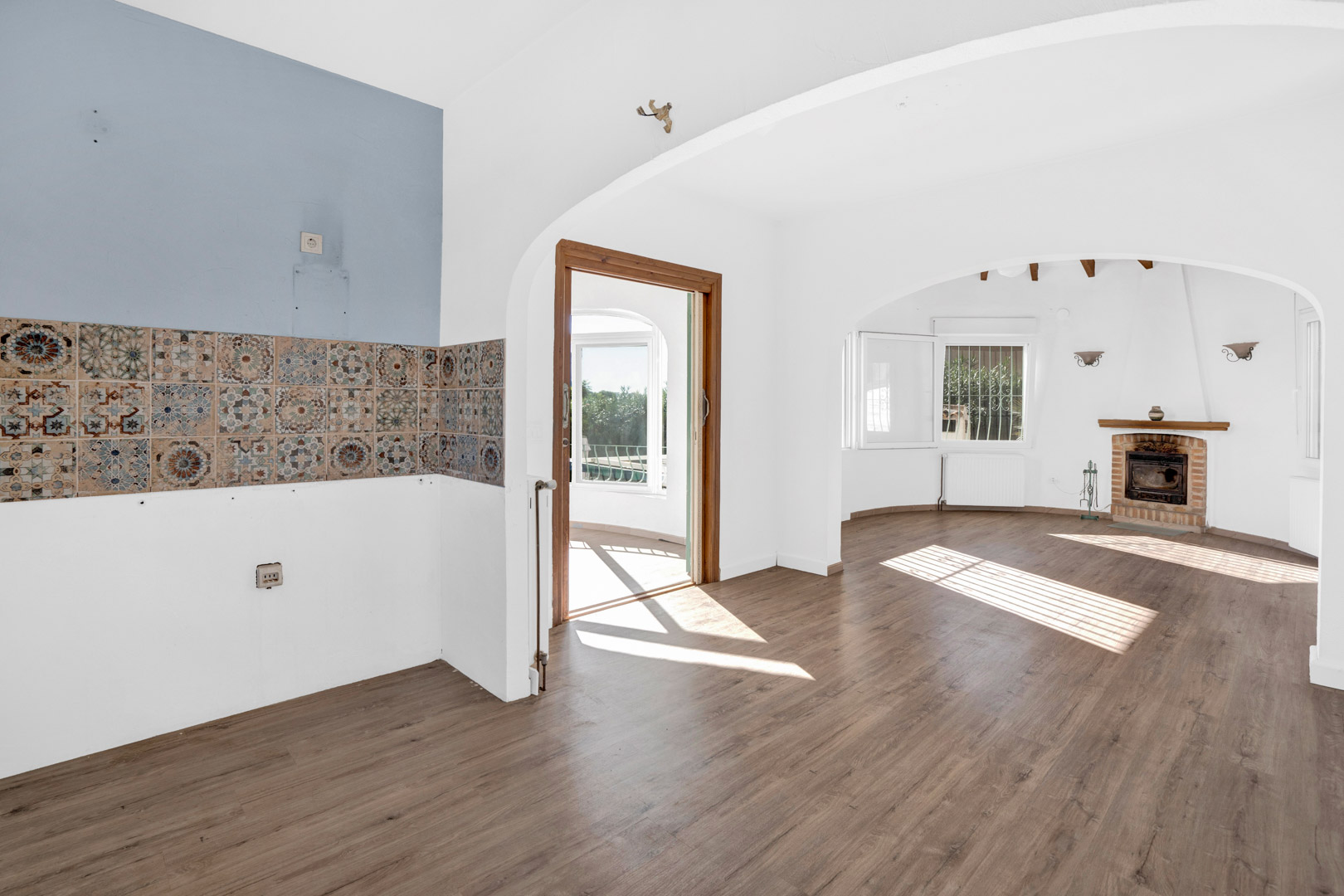 Open-plan living area featuring a brick fireplace, laminate flooring, and decorative tiled kitchen wall. Arched transitions lead to a sunlit glazed terrace.