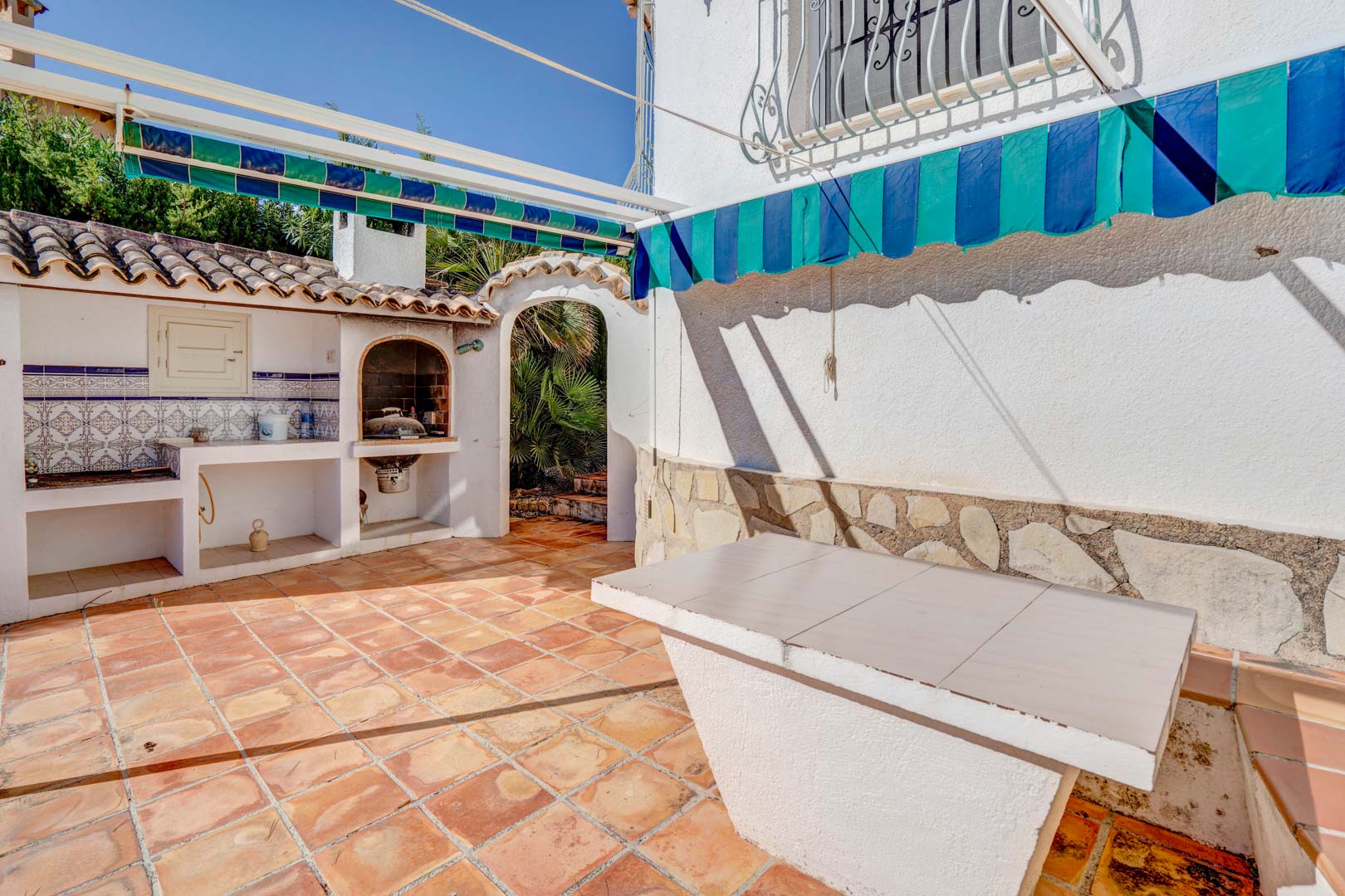 Outdoor terrace featuring a built-in barbecue with traditional tiling, a fixed stone table, and retractable awnings over terracotta flooring.