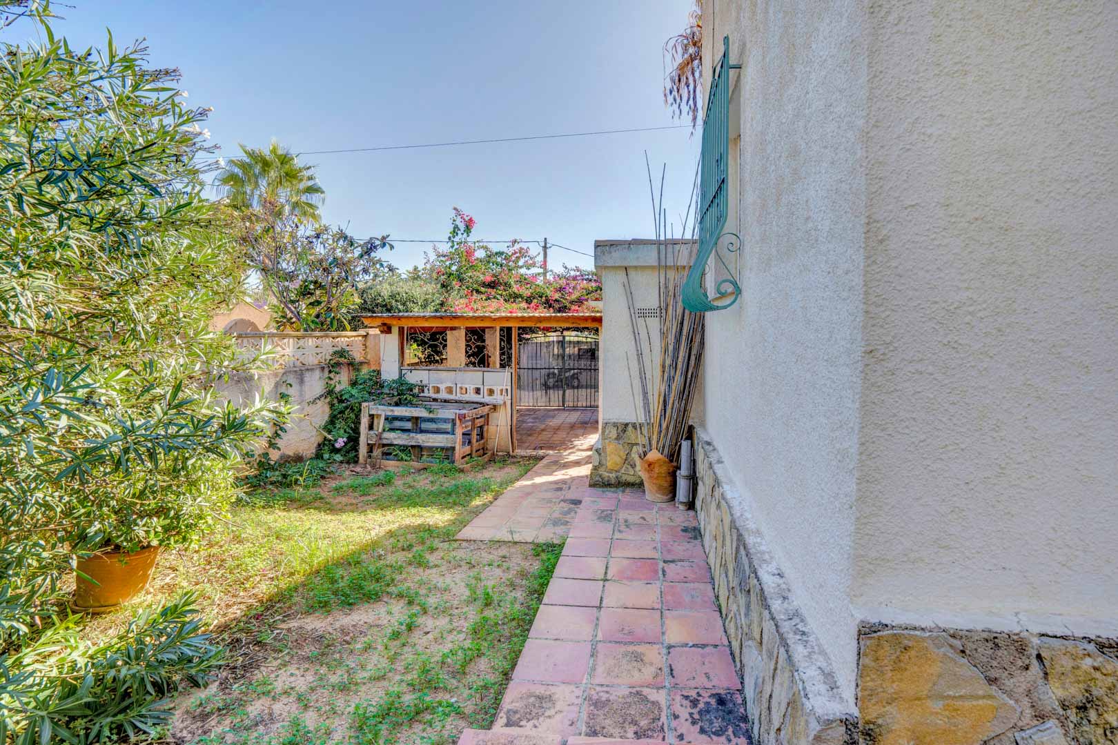 Paved side path leading to the covered carport, featuring white textured walls with a natural stone base and surrounding Mediterranean greenery.