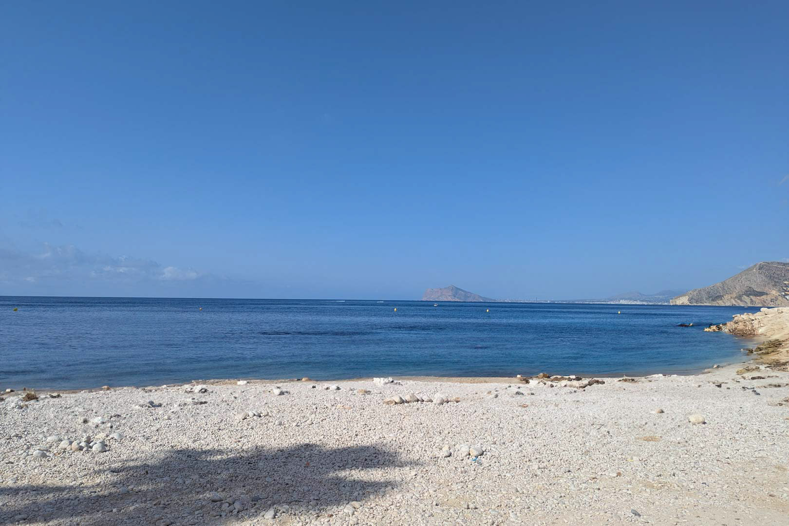 View of a white pebble cove with calm blue waters in Calpe, featuring the Mediterranean coastline and distant mountains under a clear sky.