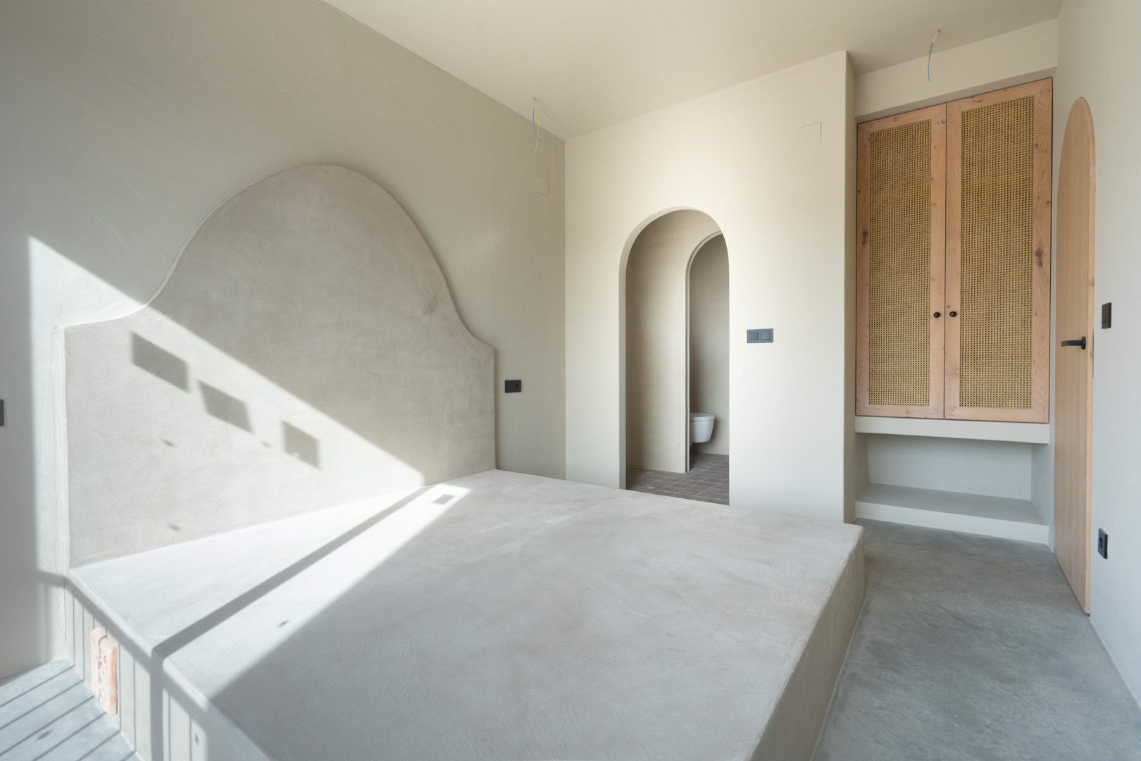 Bedroom featuring a built-in bed base with a curved clay headboard. Includes custom oak wardrobes with rattan inserts and elegant arched doorways to the ensuite.