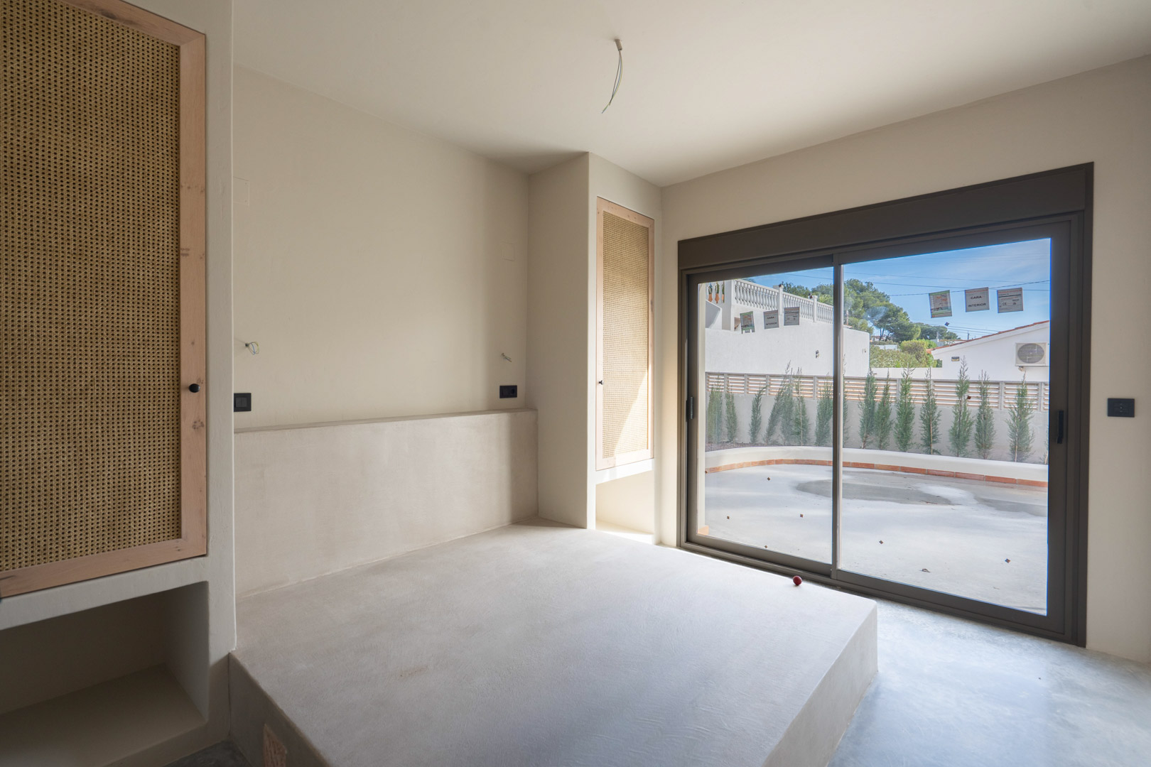 Bedroom featuring a built-in polished concrete bed platform, custom oak wardrobes with rattan inserts, and large sliding doors opening to the patio.