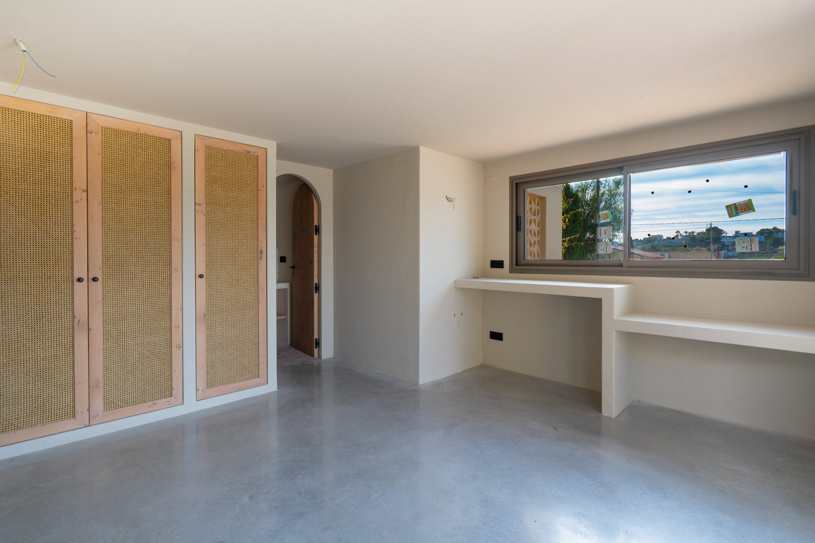 Bedroom featuring built-in wardrobes with wicker panels, polished concrete flooring, and a custom integrated desk area beneath the window and arched doorway.