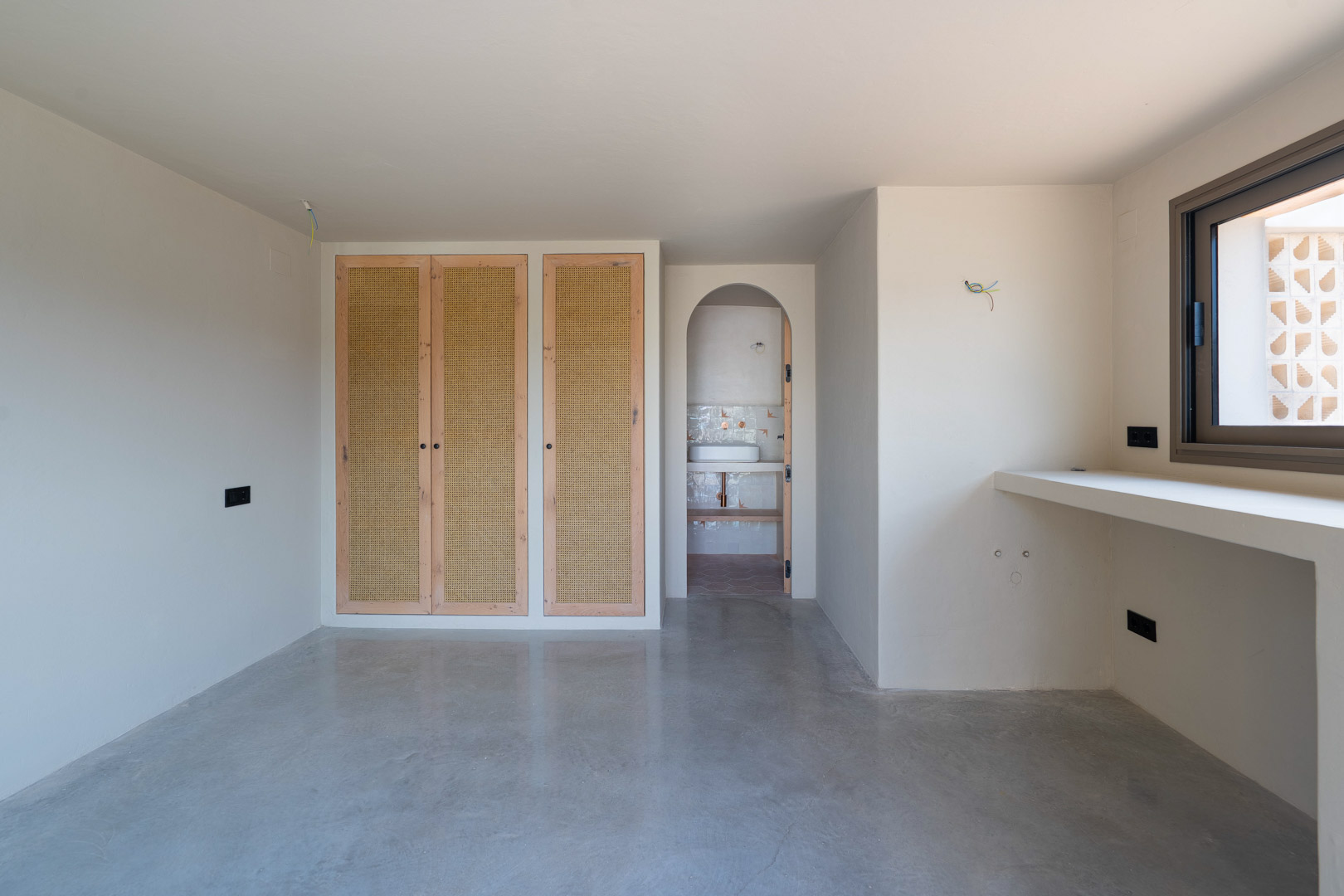 Bedroom featuring oak-framed wardrobes with rattan panels, polished concrete flooring, and an arched entrance to an en-suite bathroom.